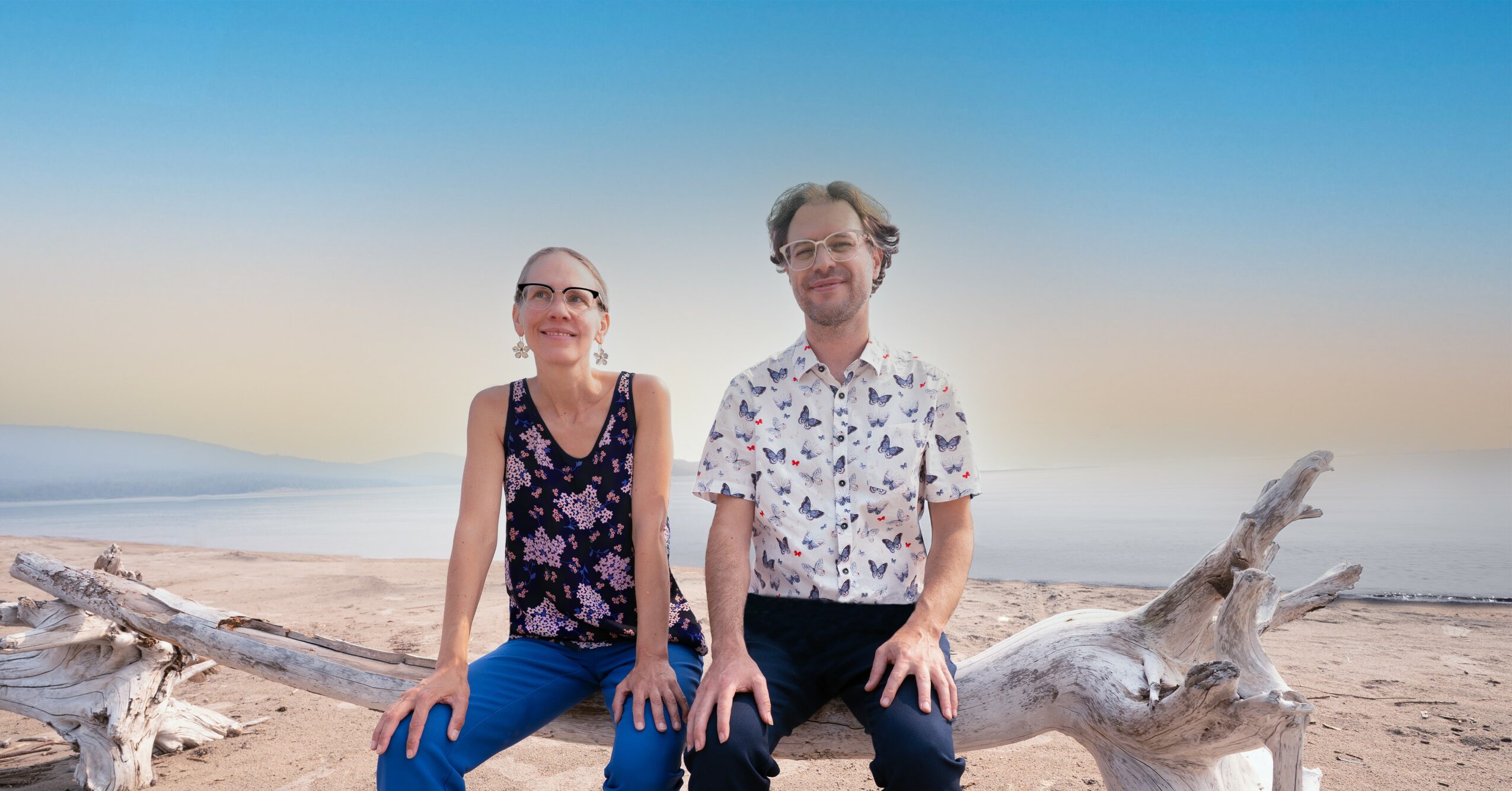 Two people sitting on driftwood on a beach with a calm sea and clear sky in the background.
