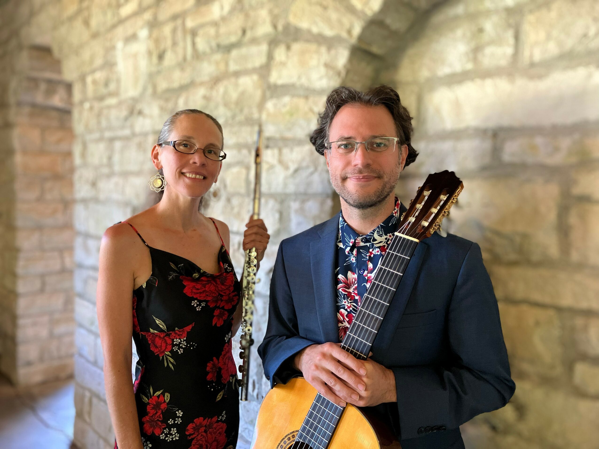 Two people standing in front of a stone wall, one holding a guitar, smiling, with a woman in a floral dress and a man in a suit.