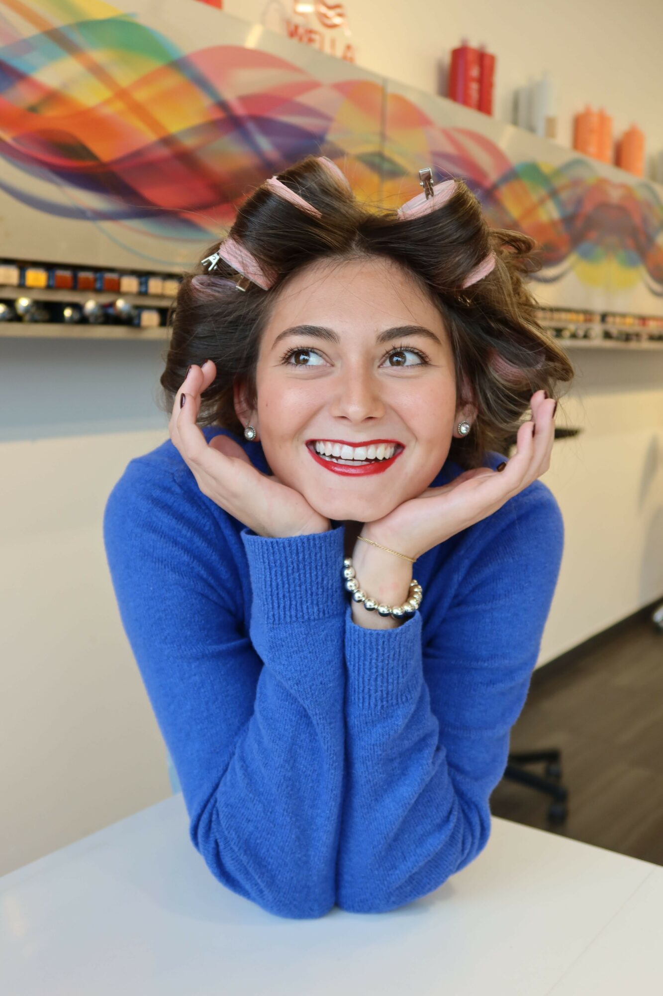 Woman with hair rollers, smiling, wearing a blue sweater, resting her head on her hands, colorful background, white table.