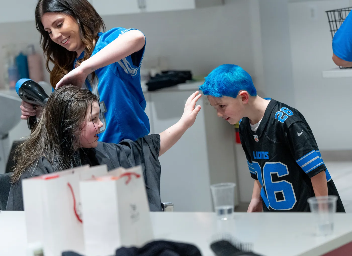 Children and a woman with blue hair in a room, children smiling and interacting, some holding objects, table with items.