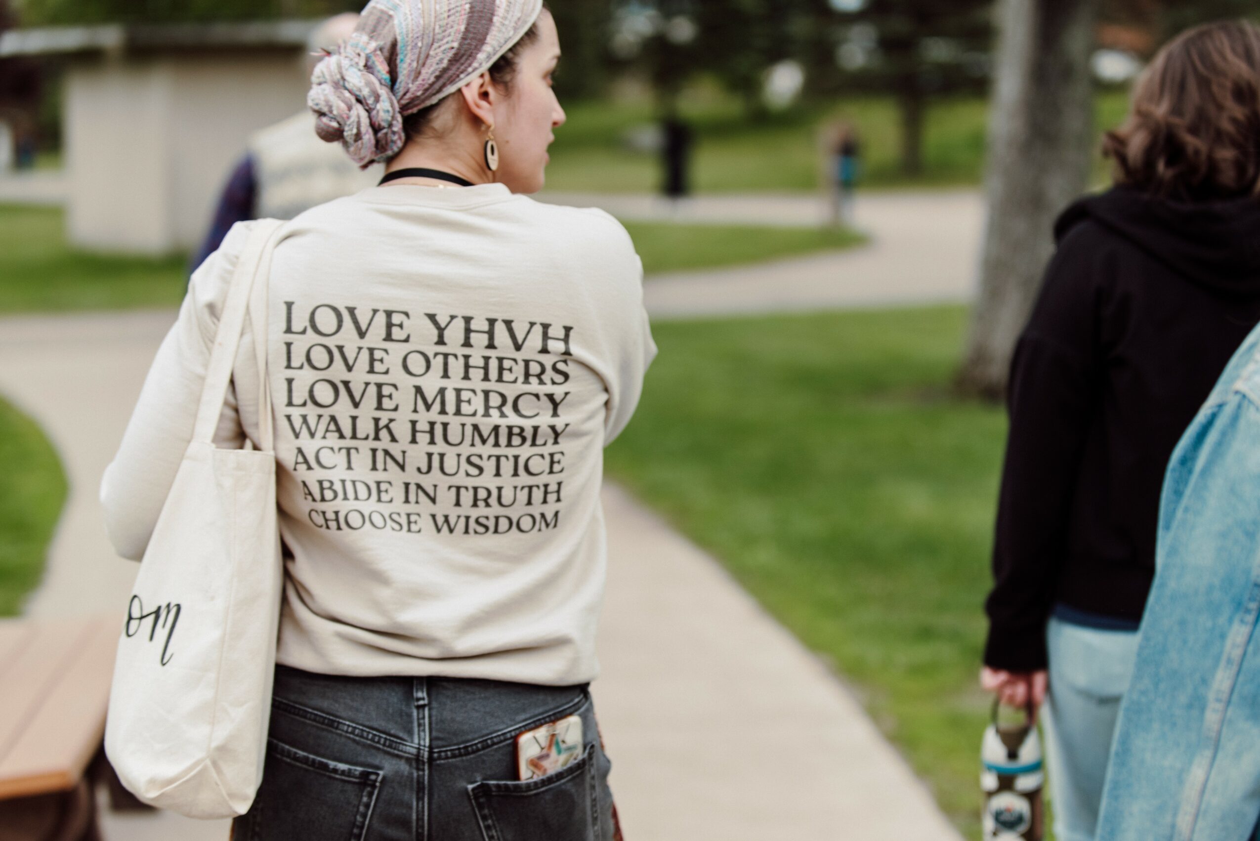 Person wearing a white sweatshirt with text, walking outdoors on a path, with trees and grass in the background.