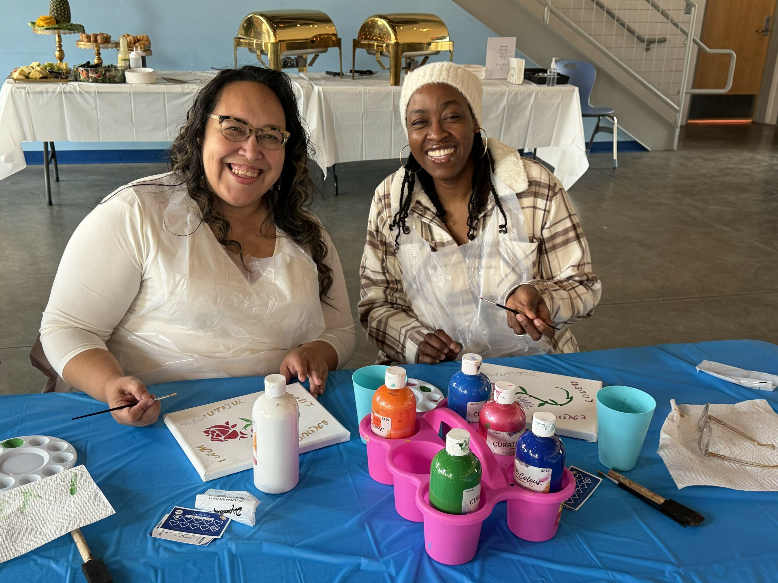 Two women sitting at a table with colorful bottles, smiling, in a room with buffet tables in the background.