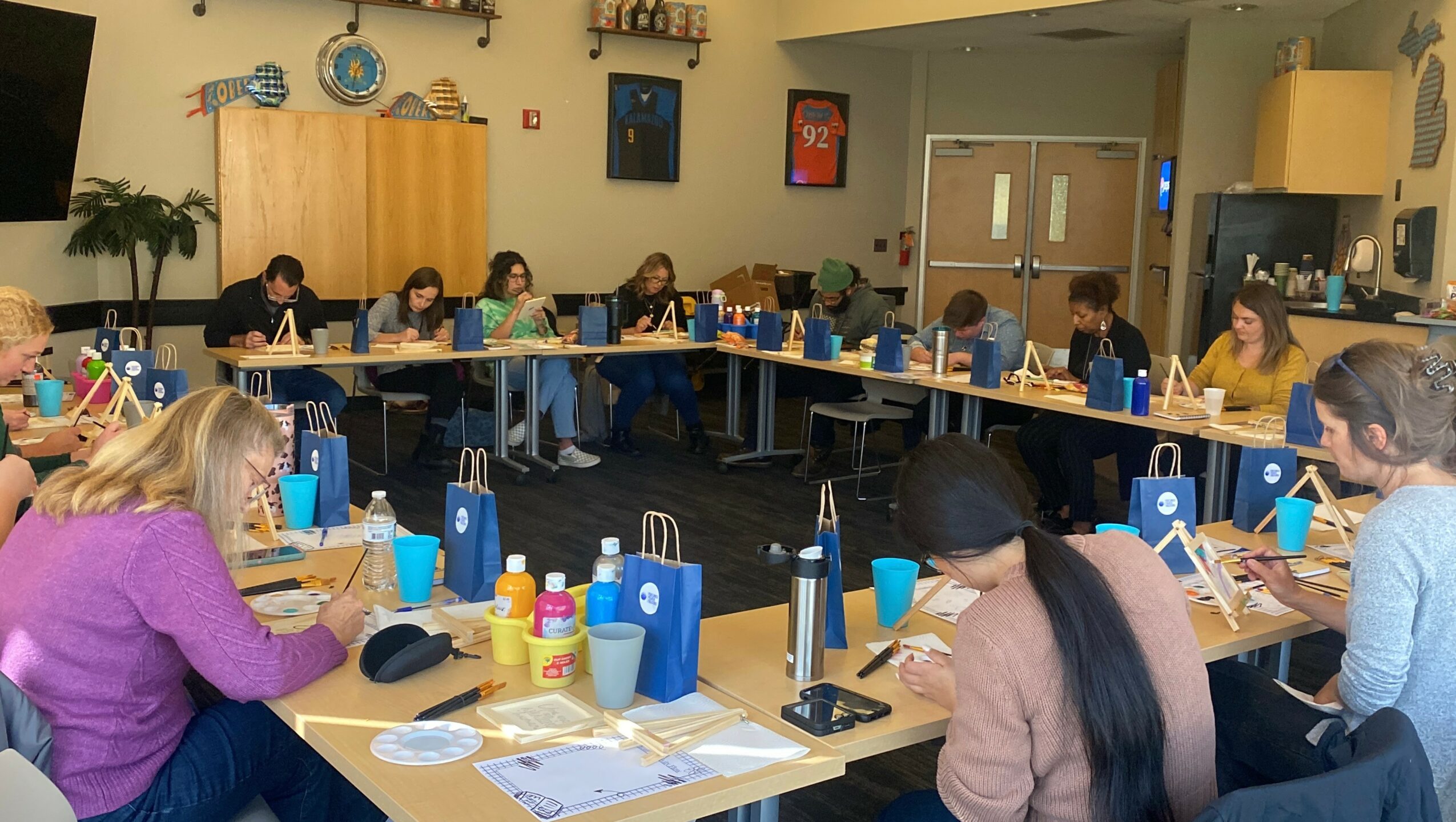 Group of people sitting around tables in a room, engaging in an activity, with supplies and blue bags on the tables.