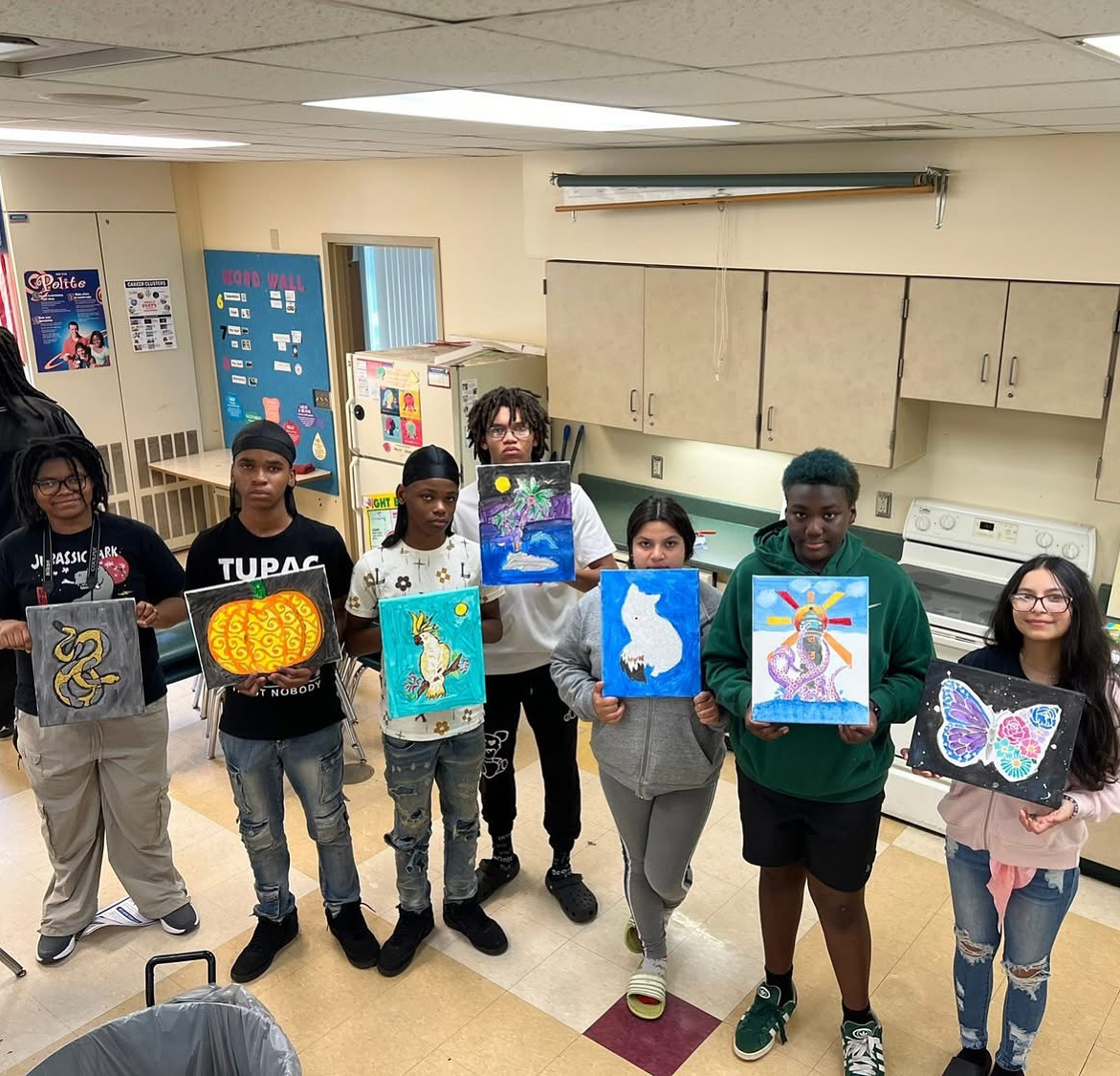Group of children holding colorful artwork in a classroom, with cabinets and posters in the background.
