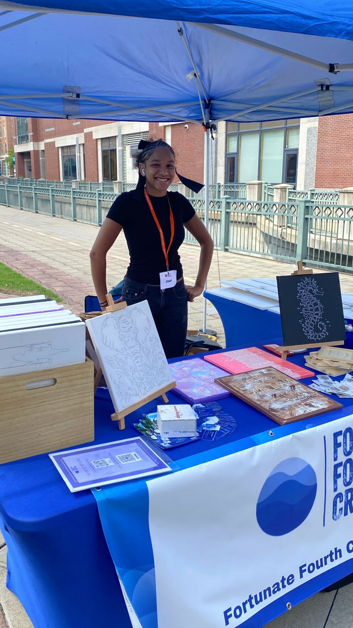 Woman standing behind a table with informational materials under a blue canopy outdoors, with buildings in the background.