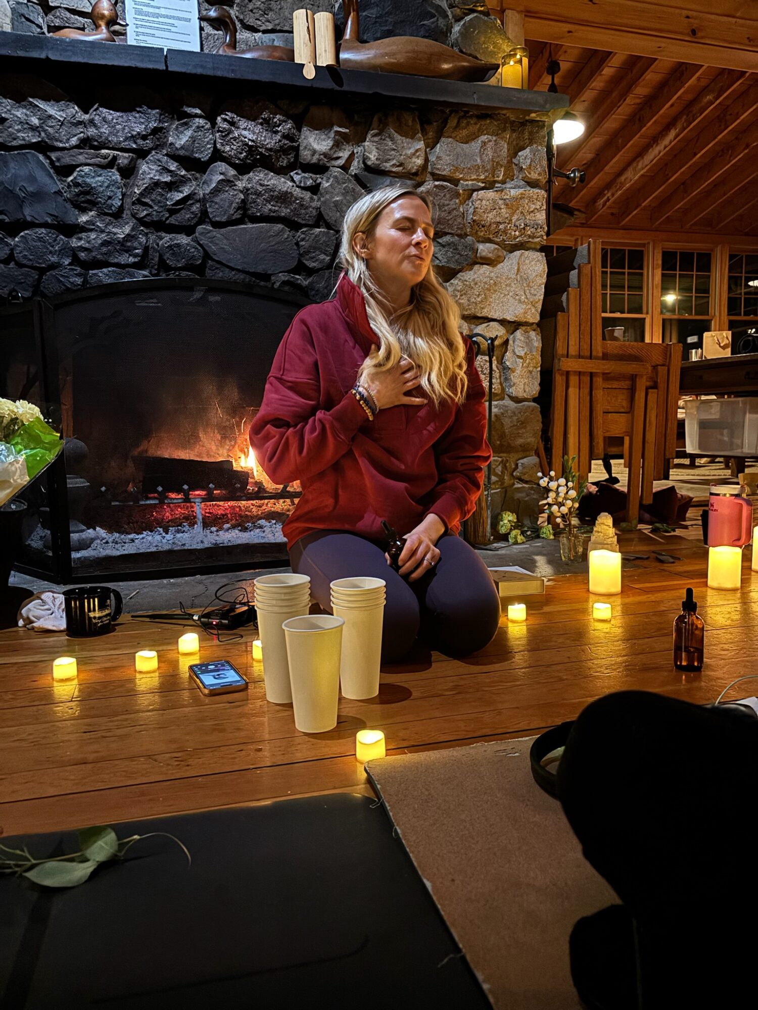Woman kneeling by a fireplace with candles around her, holding her chest, in a cozy room with wooden walls.