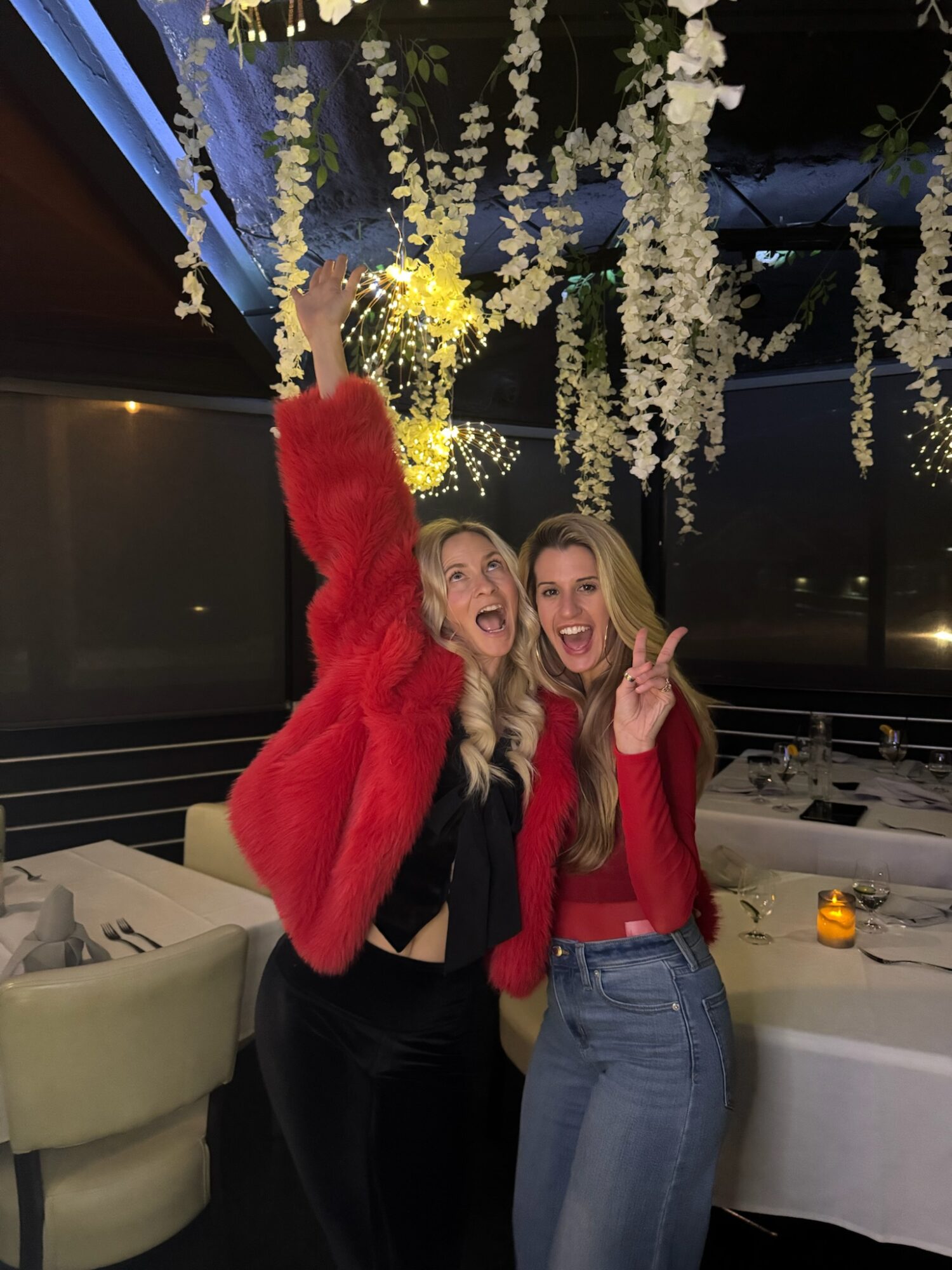 Two women smiling and making peace signs, standing under hanging white floral decorations in a decorated indoor setting.
