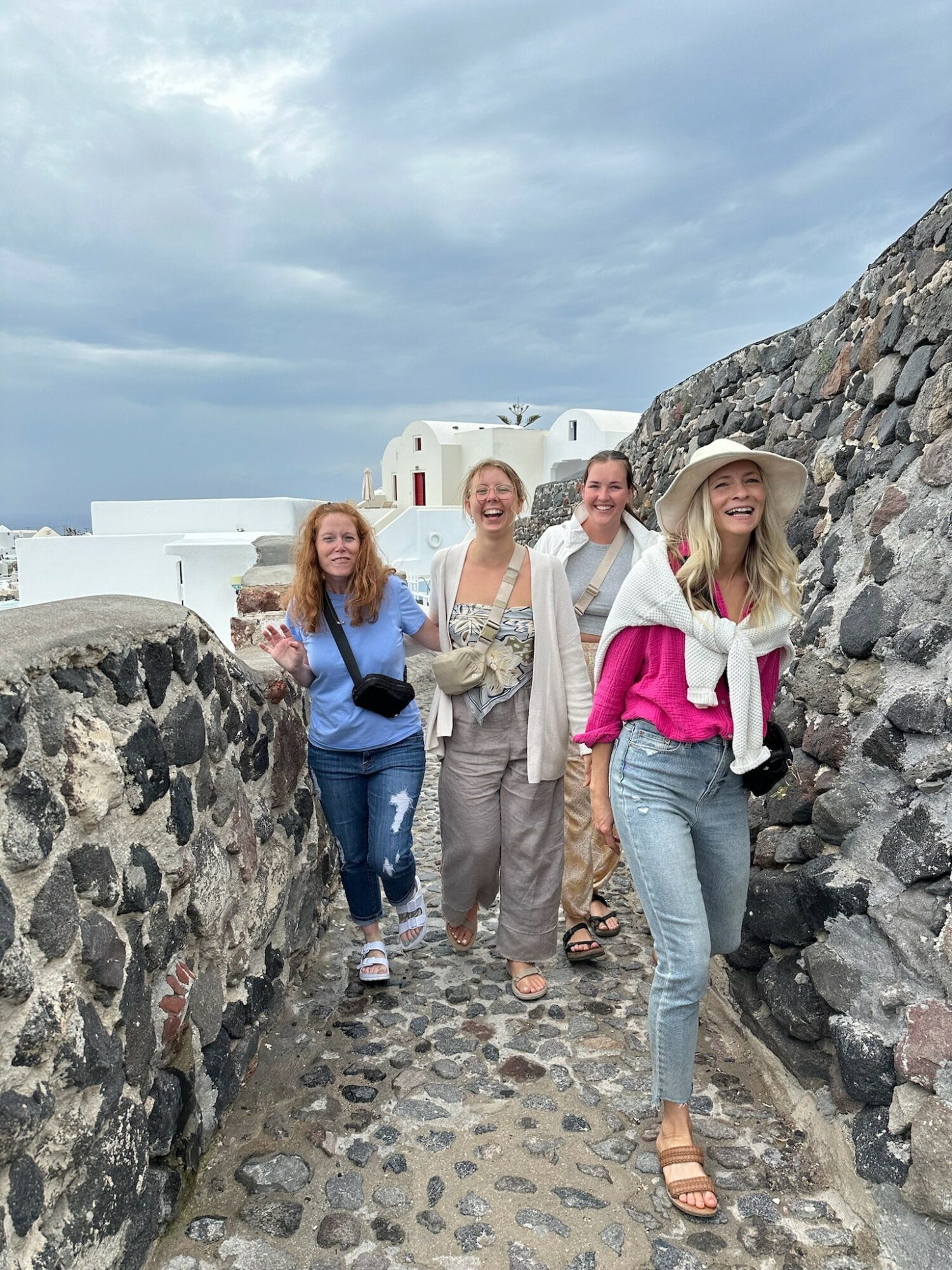 Four women walking along a stone pathway between stone walls, with white buildings and a blue sky in the background.