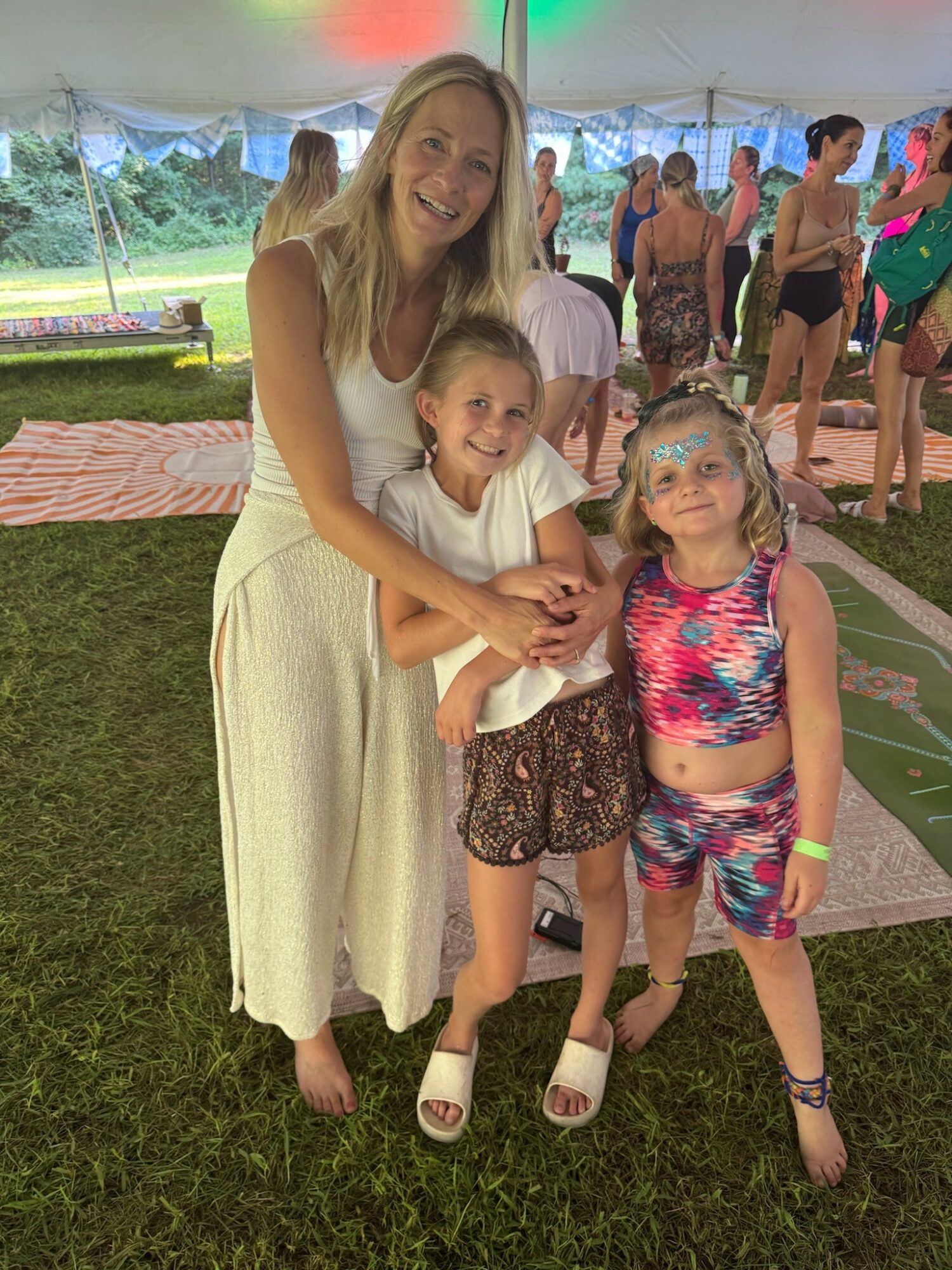 Woman hugging two children outdoors at a gathering, with people in the background under a tent, grass, and trees.