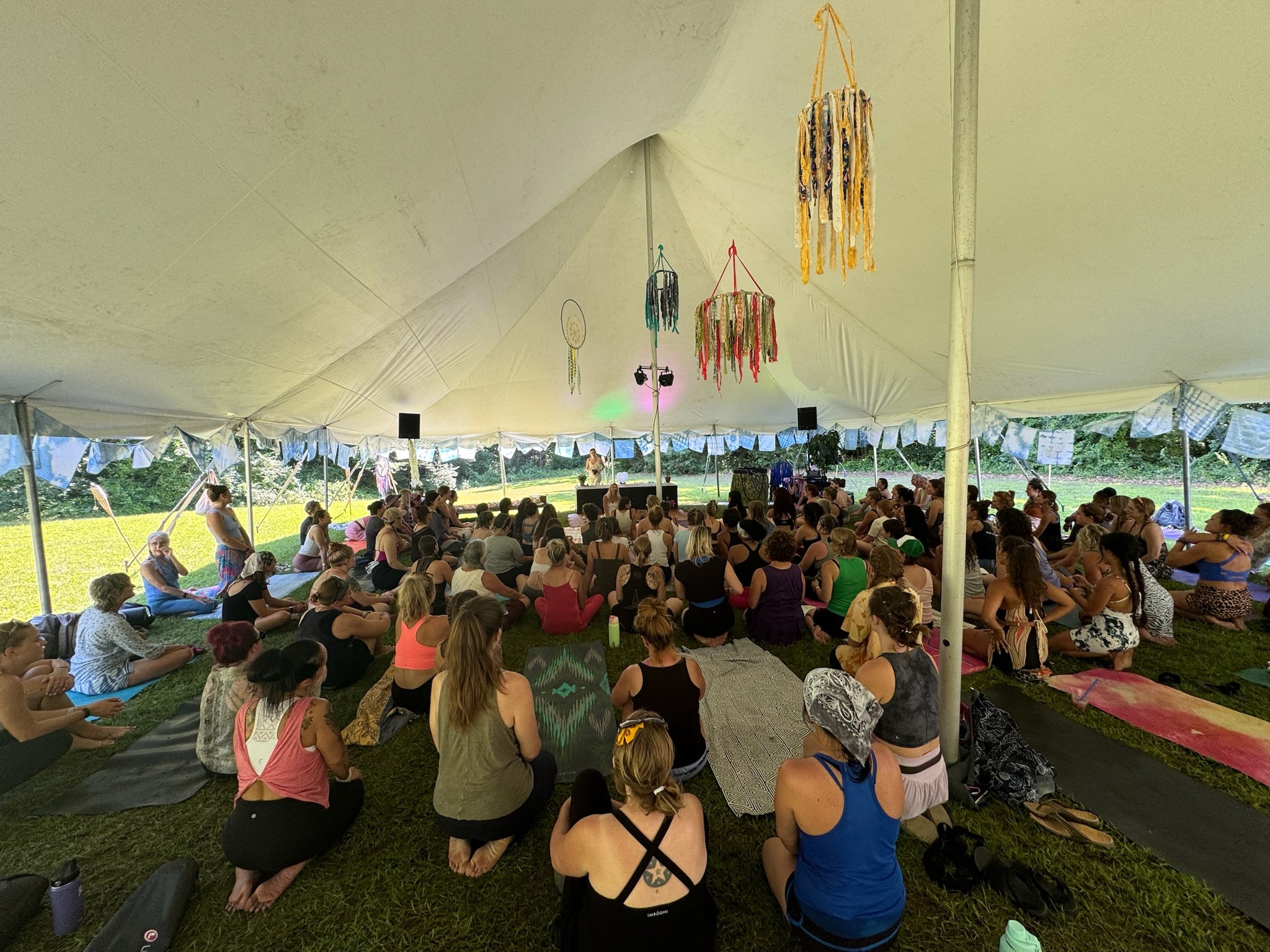 People sitting on mats inside a large tent, facing a stage with hanging decorations and a speaker, outdoors.