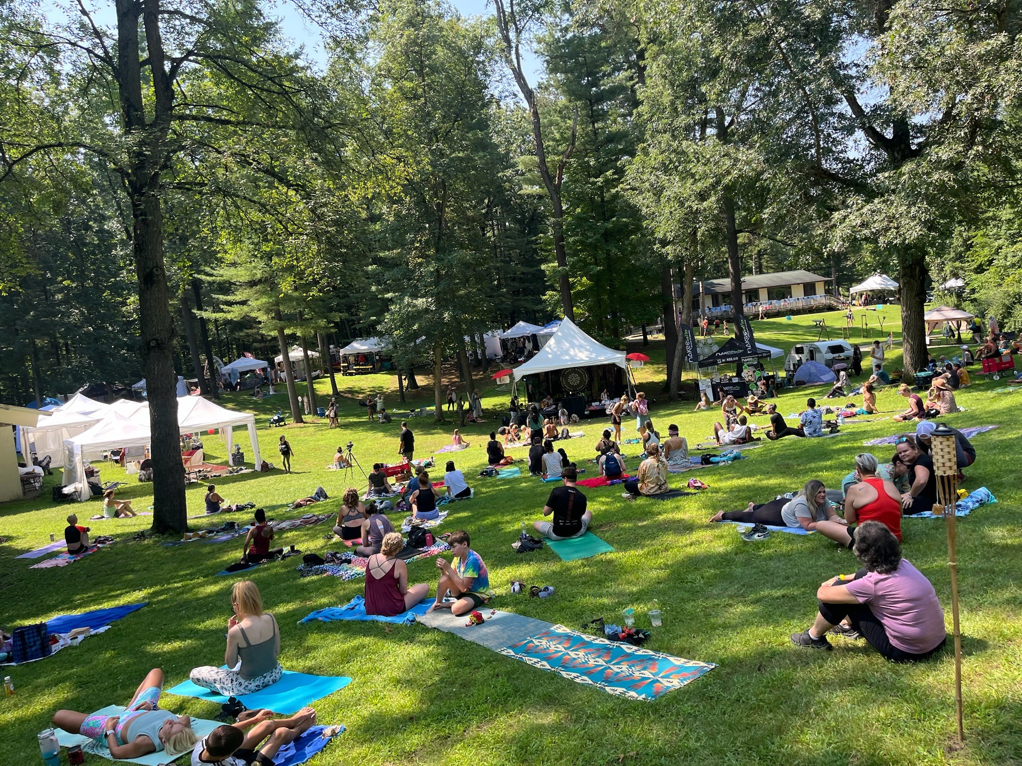 People sitting on blankets and chairs in a park with trees, tents, and a building in the background.