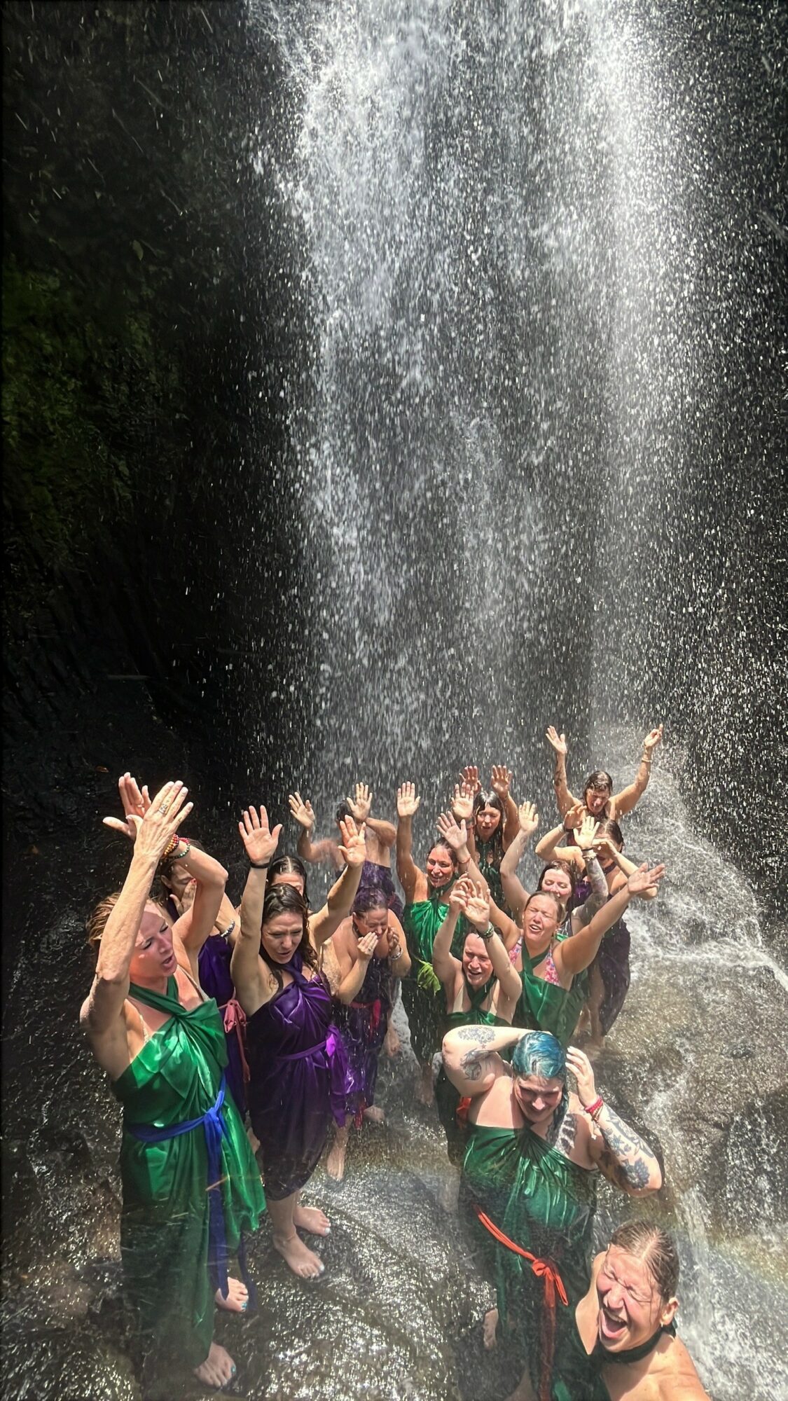 Group of people under a waterfall, raising hands and smiling, with water cascading down behind them.