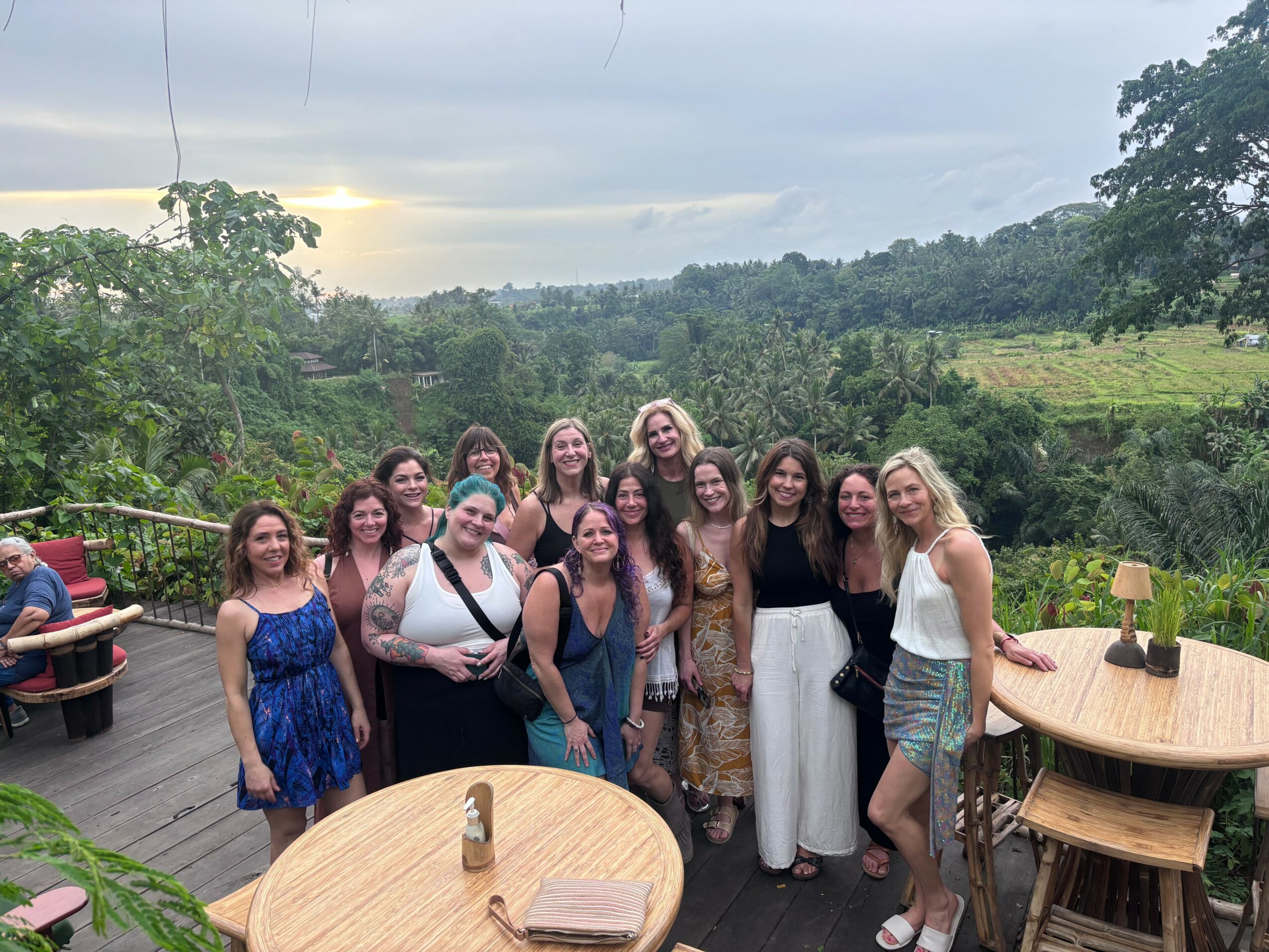 Group of ten women standing outdoors on a wooden deck with trees and hills in the background, some smiling and posing for the photo.