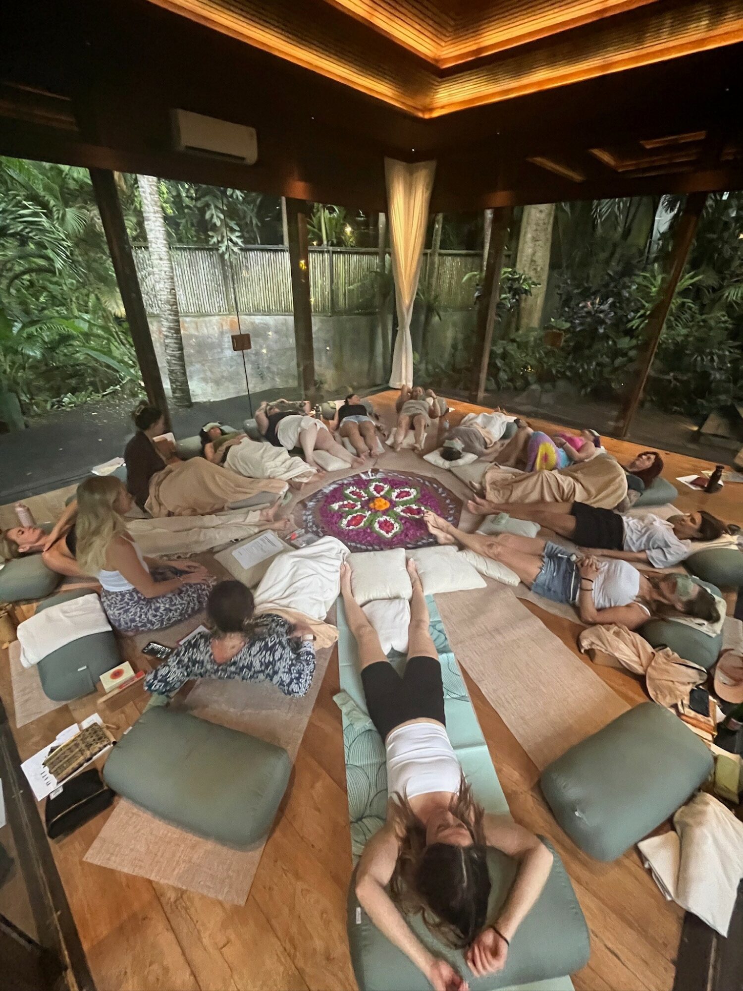 Group of people lying on cushions and mats in a circle inside a room with large windows and wooden ceiling.