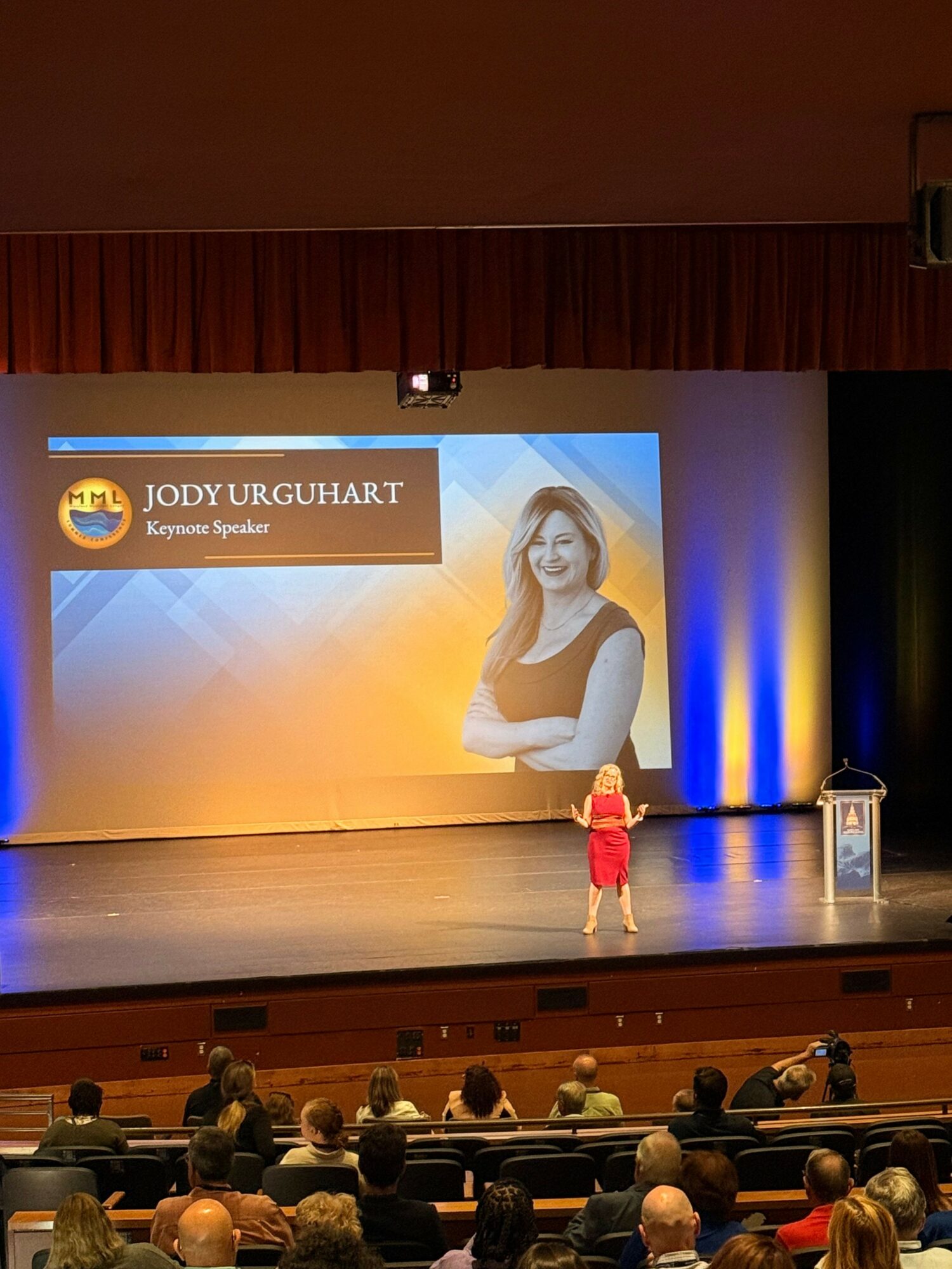 Person on stage giving a presentation in front of a large audience, with a projected slide behind them.