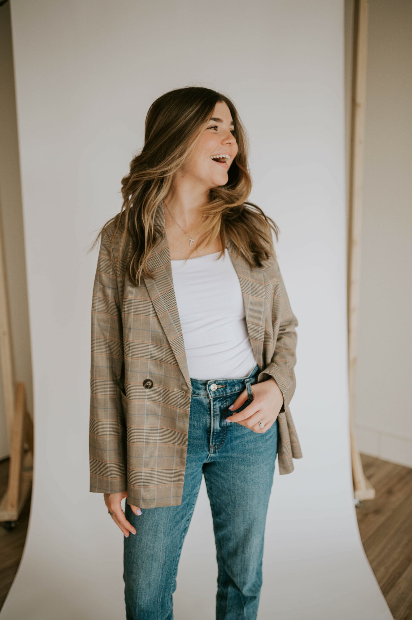Woman with long hair wearing a plaid blazer and white shirt, smiling, standing indoors against a white background.