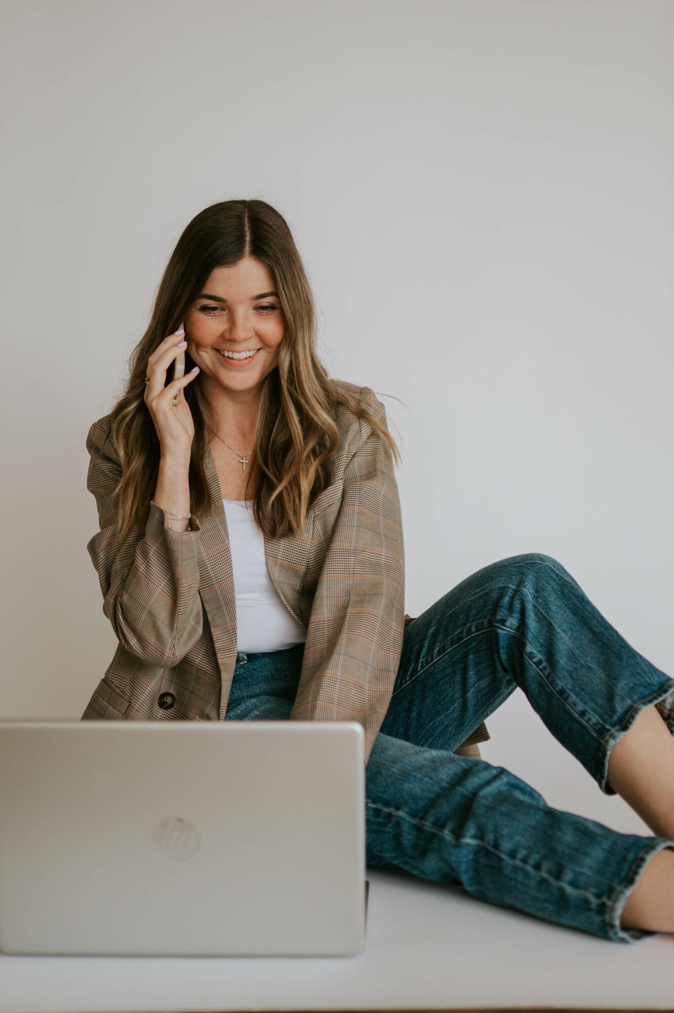 Young woman sitting on a surface, smiling, talking on phone, with a laptop in front of her, against a plain background.