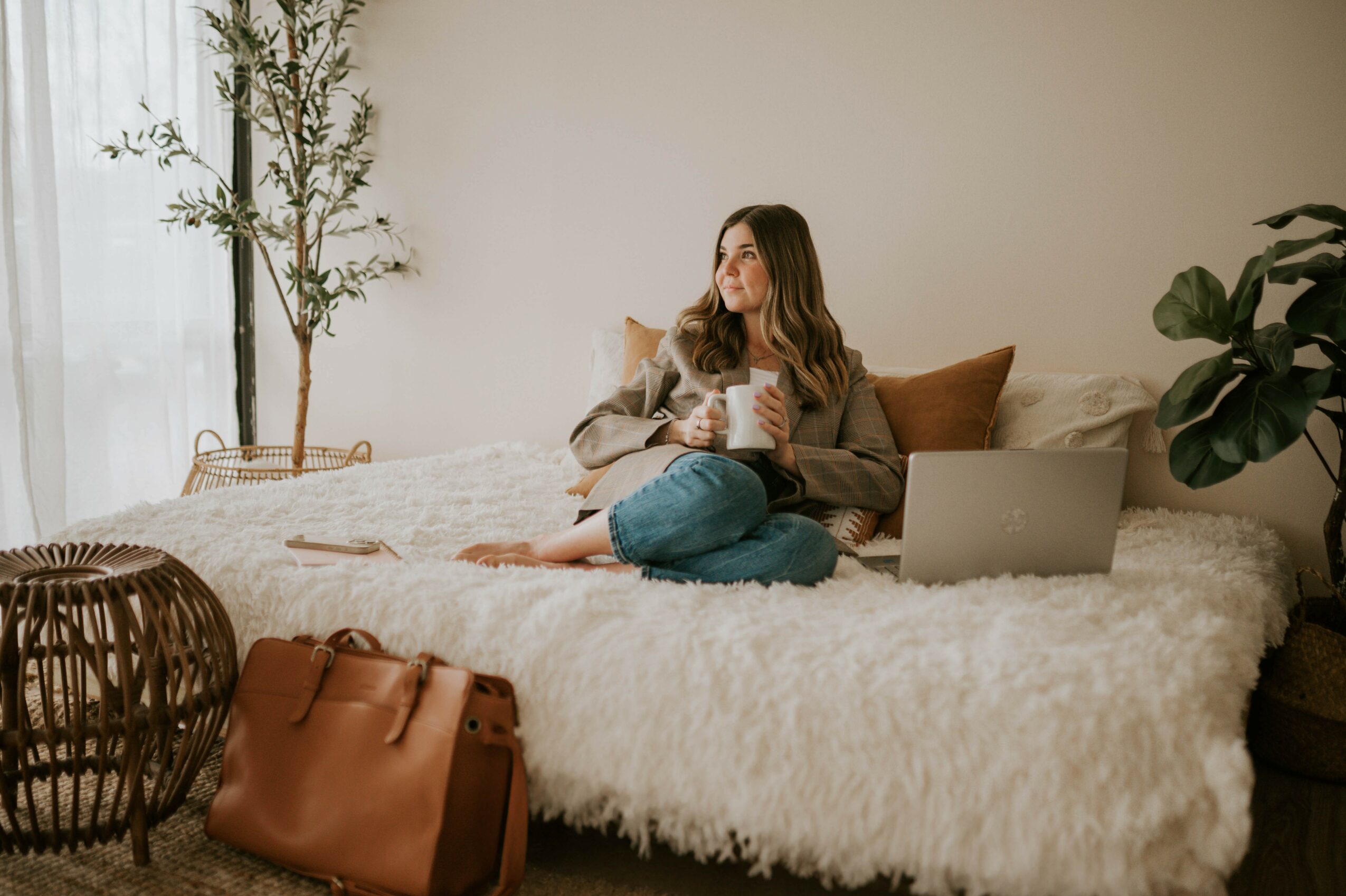 Woman sitting on a fluffy bed with a laptop and coffee, in a cozy room with plants and natural light.