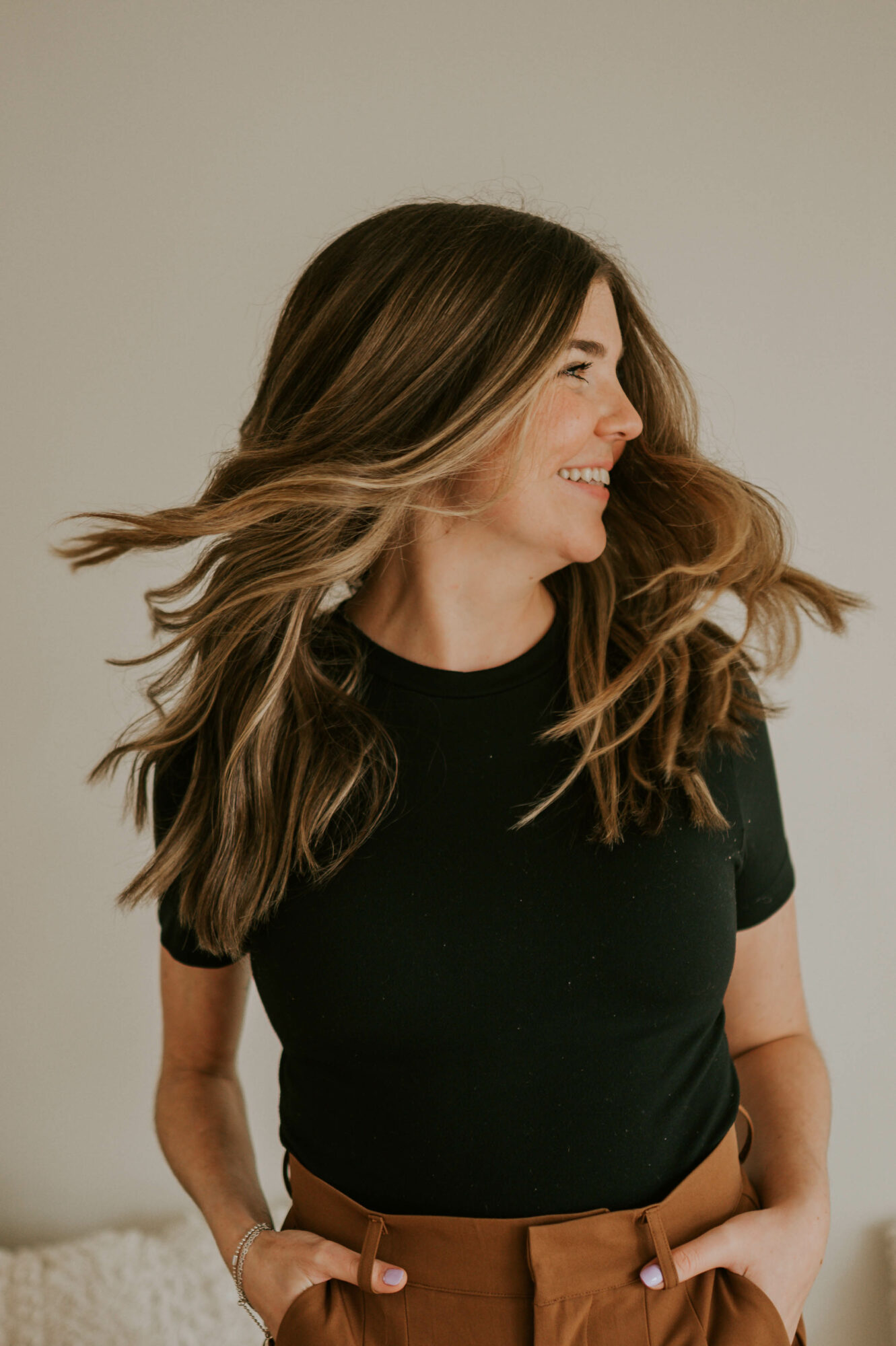 Woman with long, wavy hair smiling, wearing a black top and brown pants, facing right.
