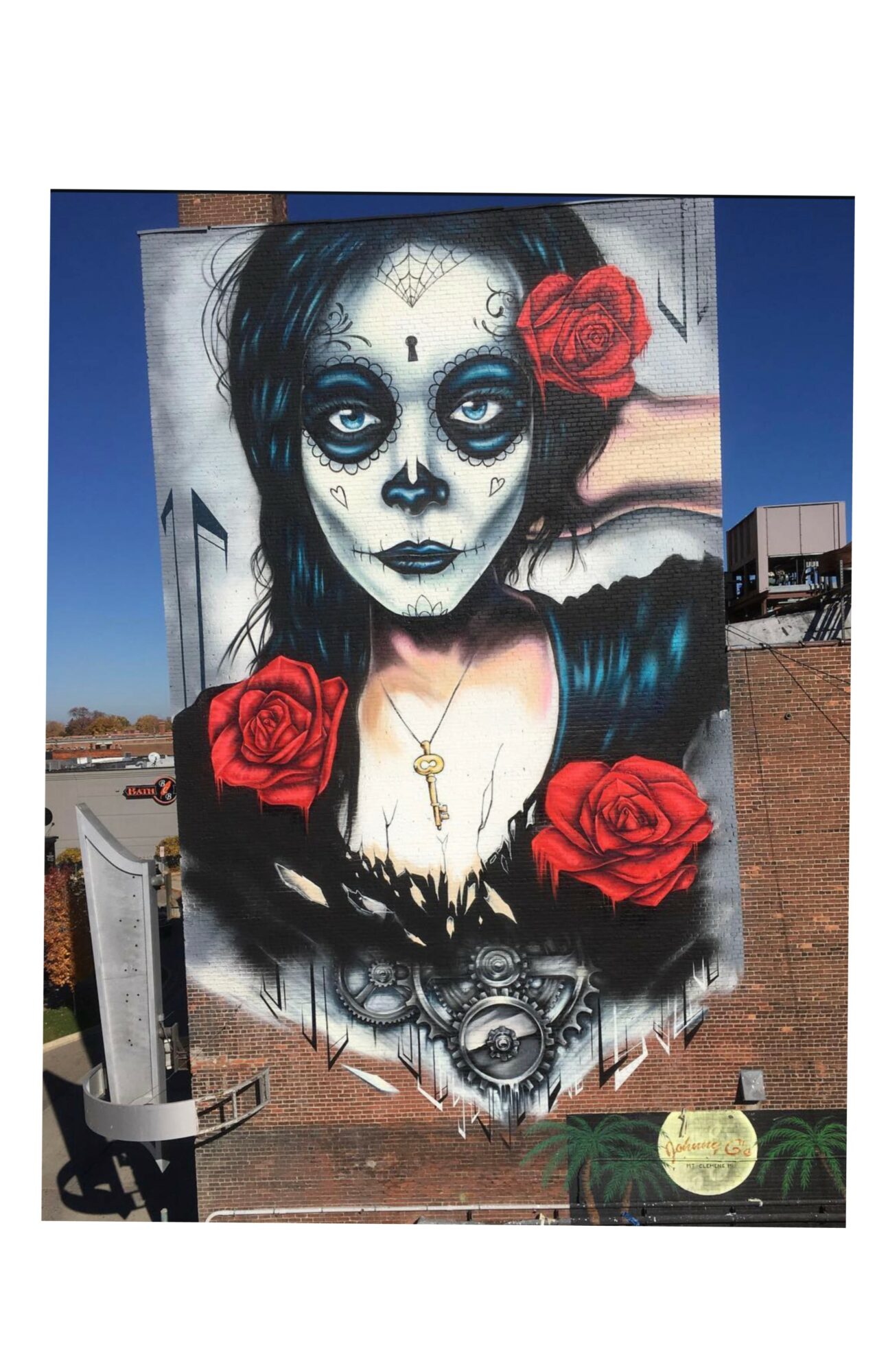 Street mural of a woman with sugar skull makeup, red roses, and dark hair, with a cityscape background.