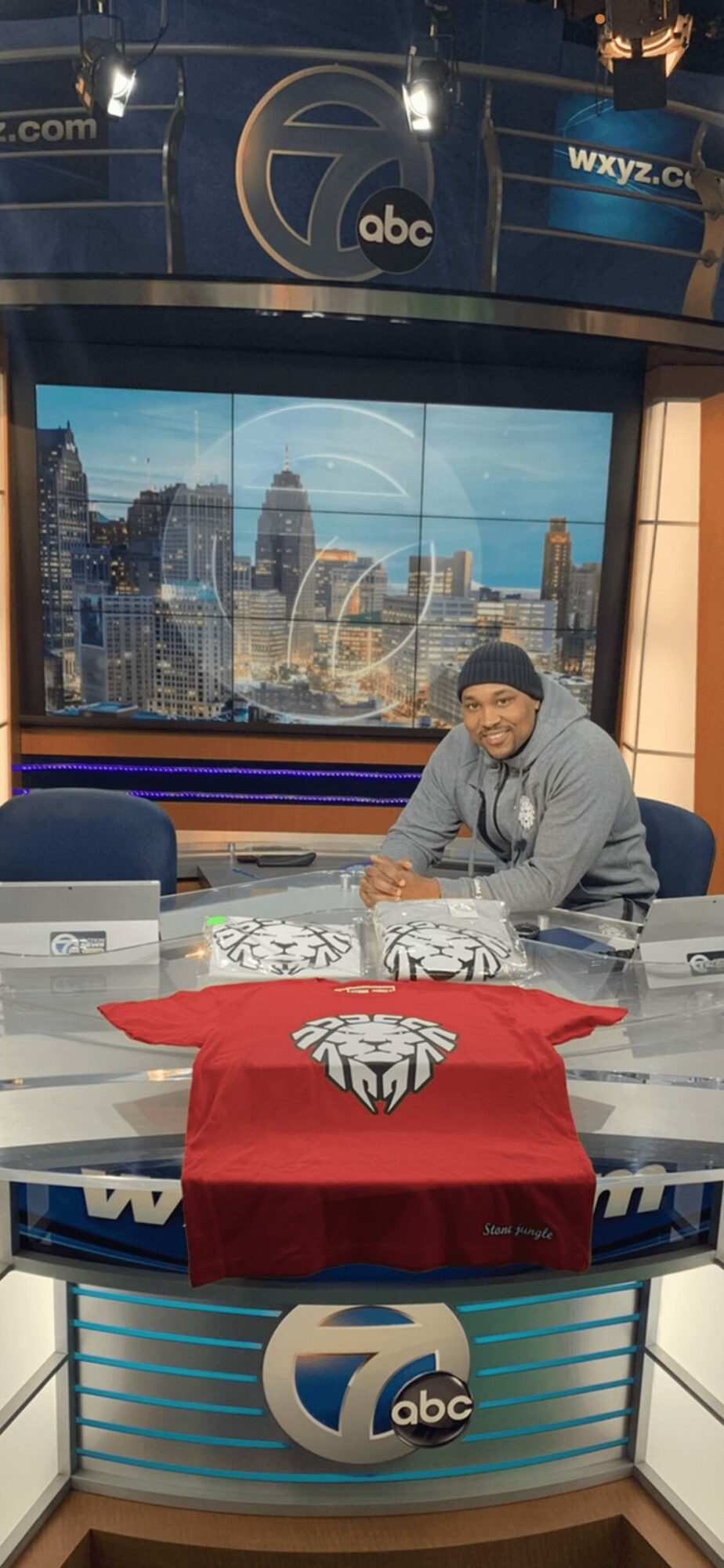 Person sitting at a news desk with a cityscape background on a large screen behind them.