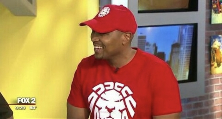 Man wearing a red cap and red shirt smiling, television screens in background, yellow wall on the left.