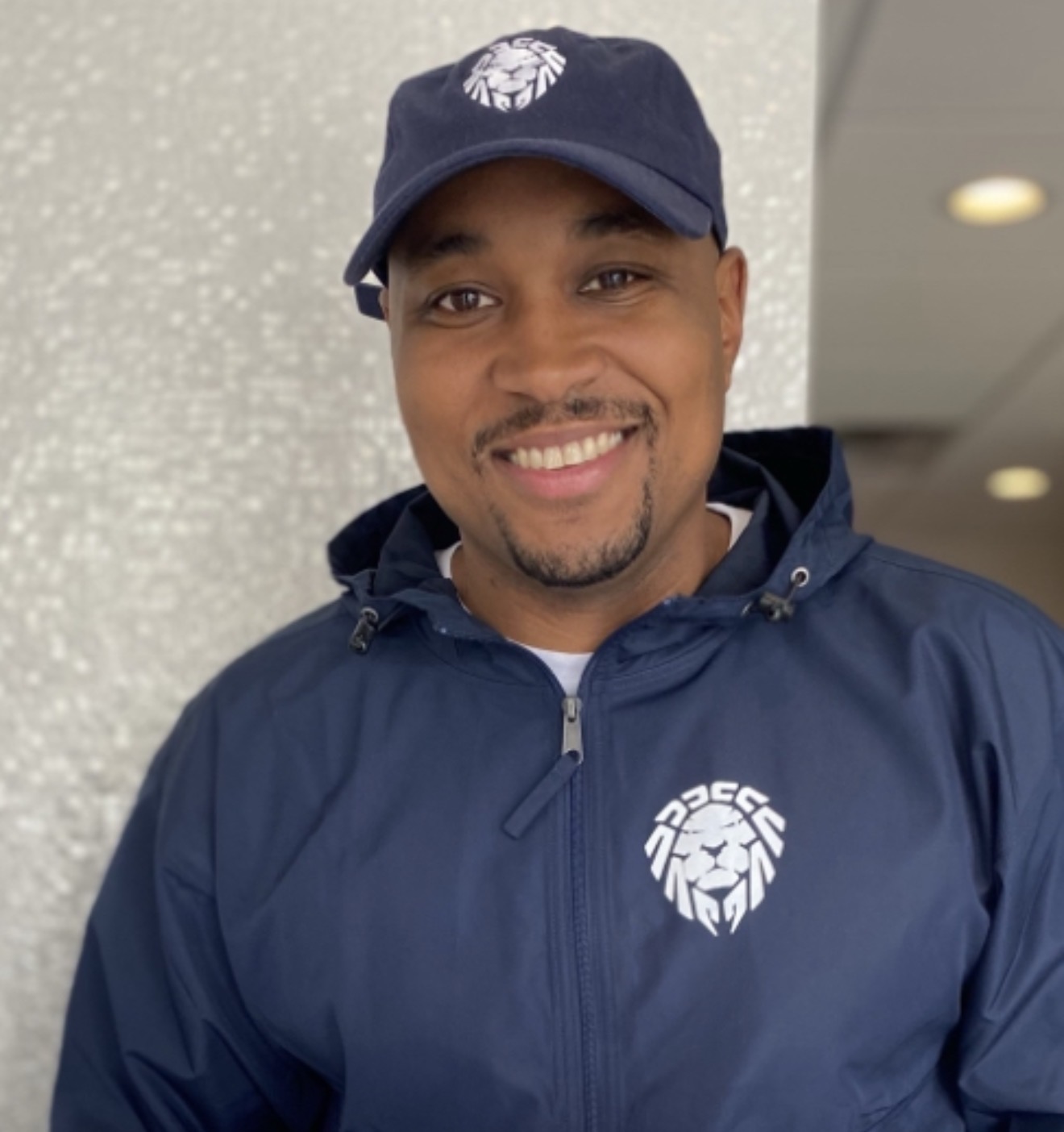 Smiling man wearing a navy blue jacket and cap with a lion logo, standing indoors against a light background.