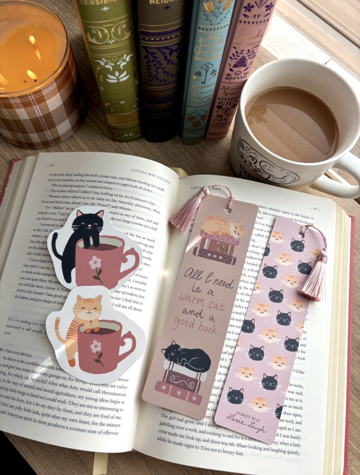 Open book with illustrations of cats and teapots, pink bookmarks, a cup of tea, and a candle on a windowsill.
