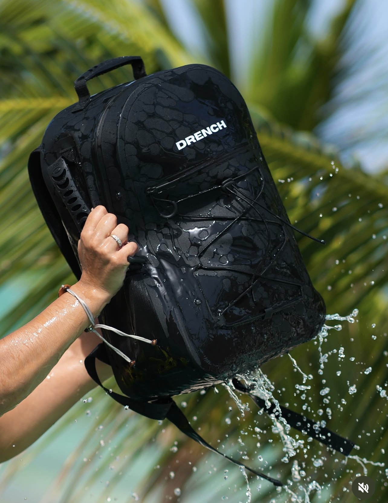 Person holding a black Drench backpack outdoors with water splashing, green plants in background.