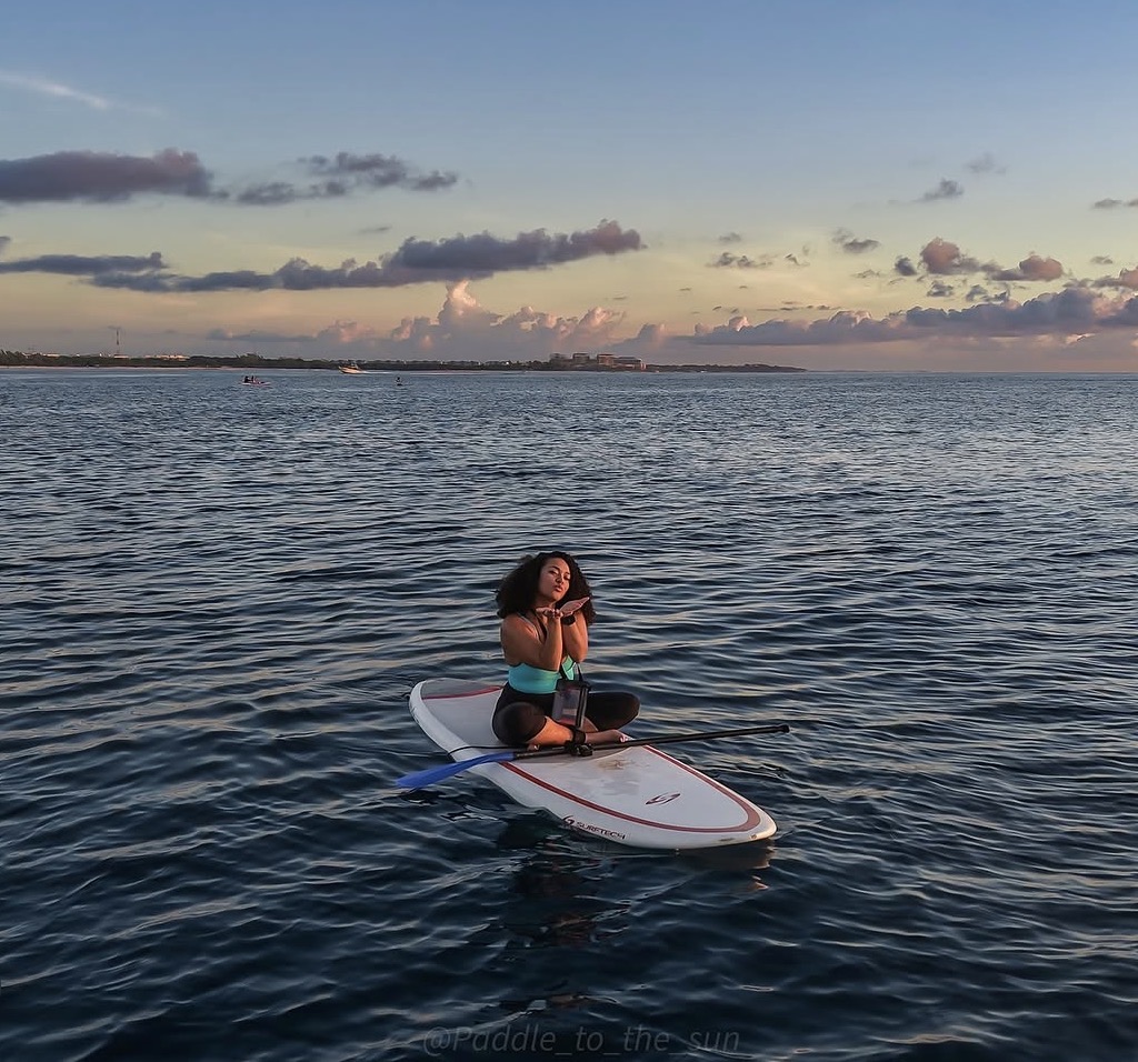 Person sitting on paddleboard on water during sunset, holding paddle, with sky and clouds in background.