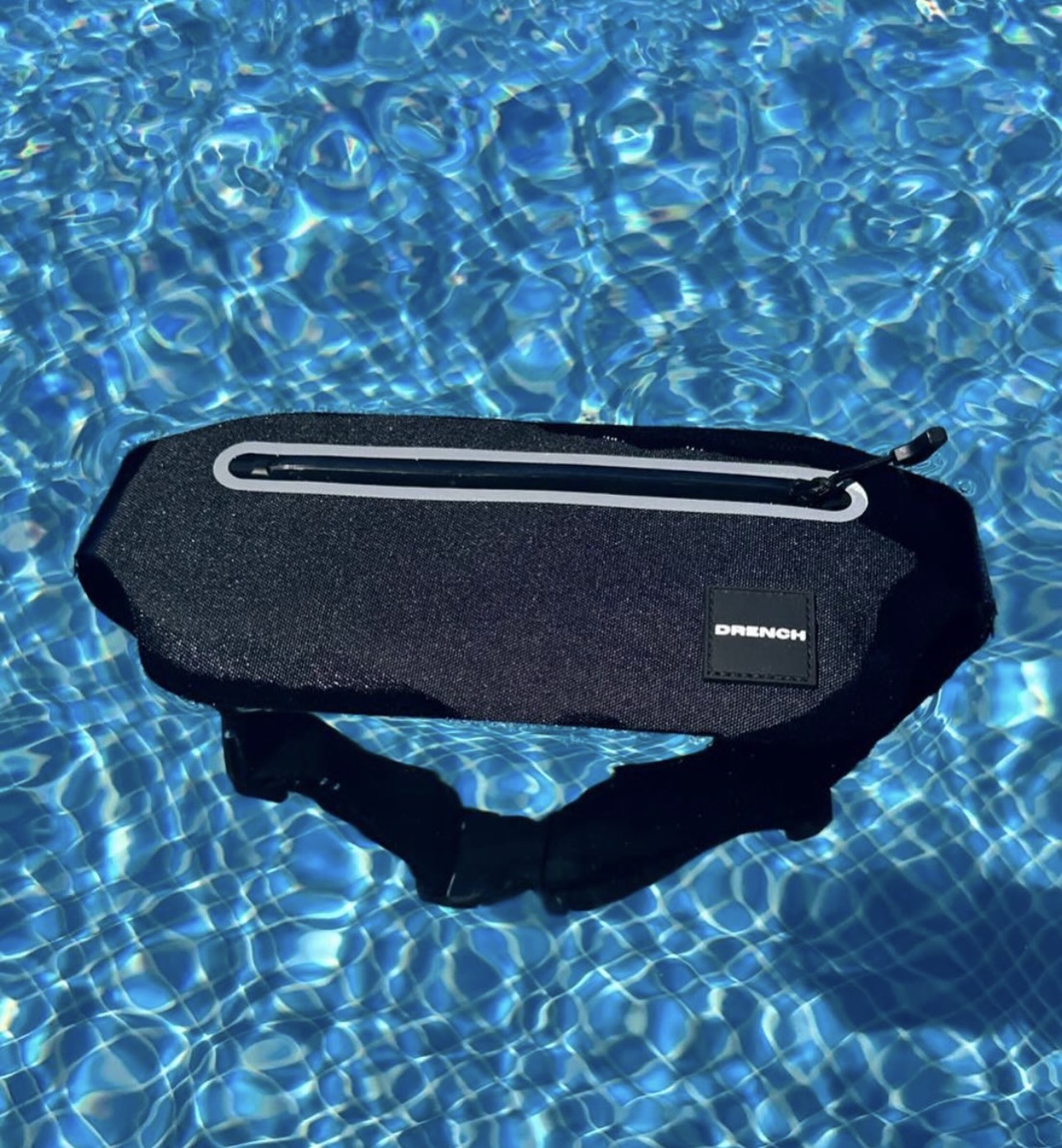Black waist bag floating on a blue water surface with sunlight reflections, zipper visible, brand label on the bag, 20 words