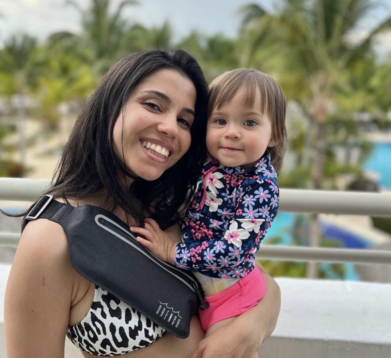 Smiling woman holding a young girl outdoors with trees and a pool in the background.