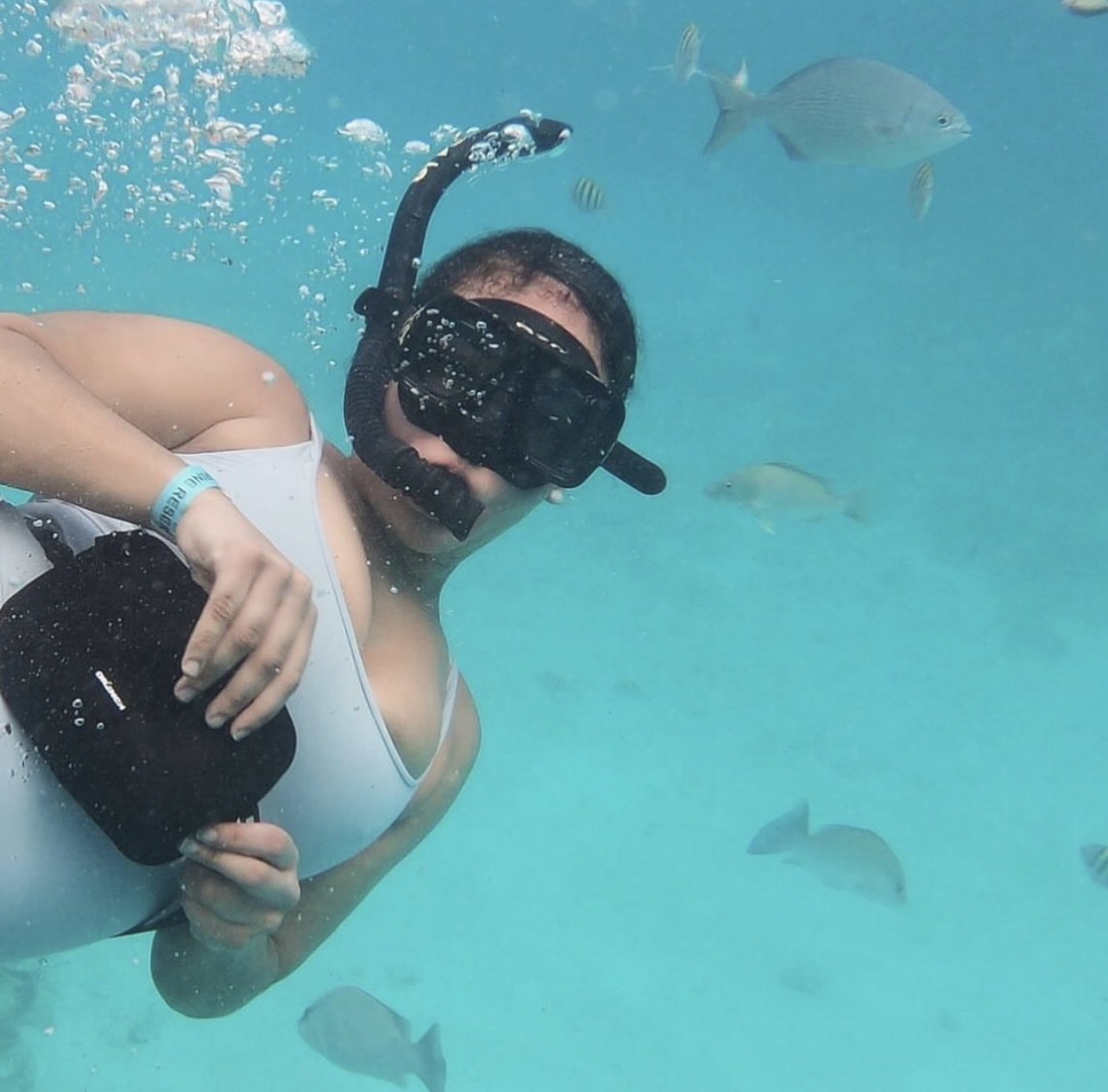 Person snorkeling underwater with fish swimming nearby, wearing mask and snorkel, holding a black object, in clear blue water.