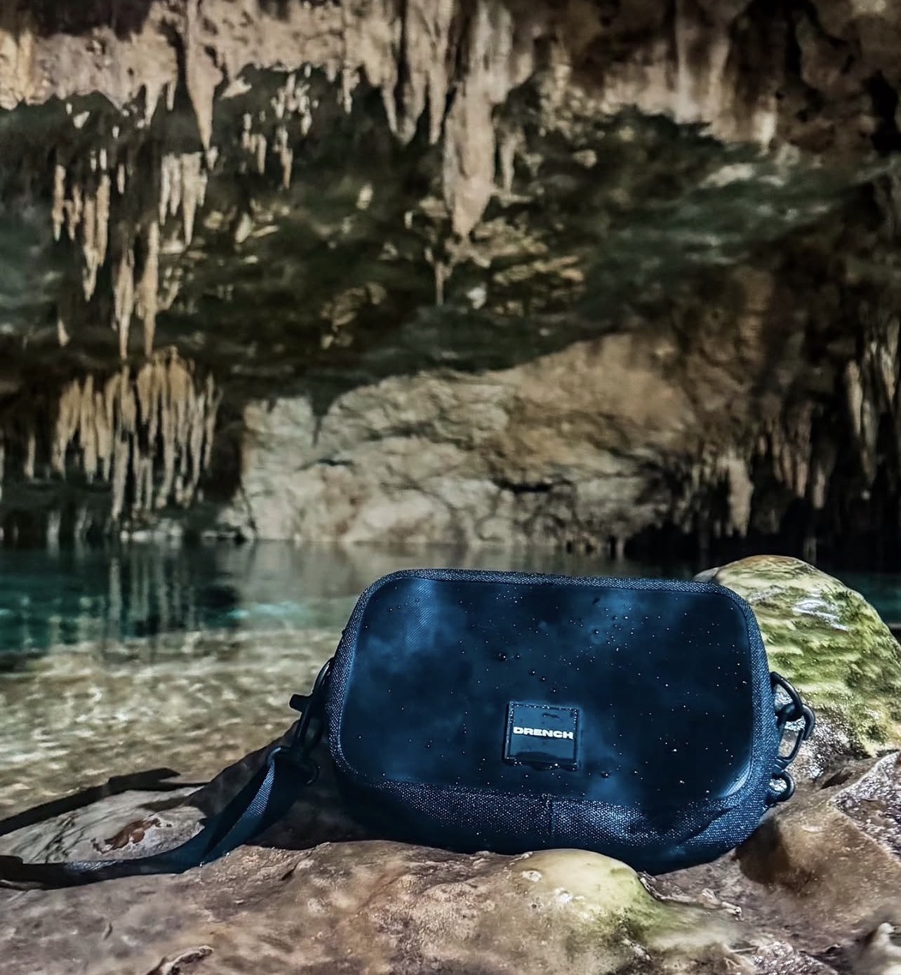 A black bag resting on rocks inside a cave with stalactites and water in the background.