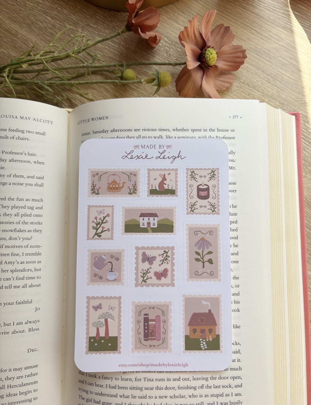 Open book on a wooden surface with a floral bookmark and pink flowers nearby, showing a sticker sheet with various designs.