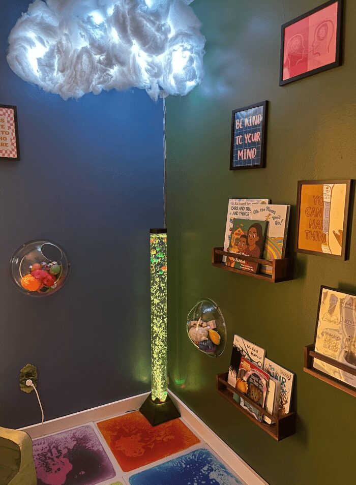 Corner of a room with a cloud-shaped ceiling light, green wall, and dark blue wall. Shelves with books and framed art, colorful floor tiles.