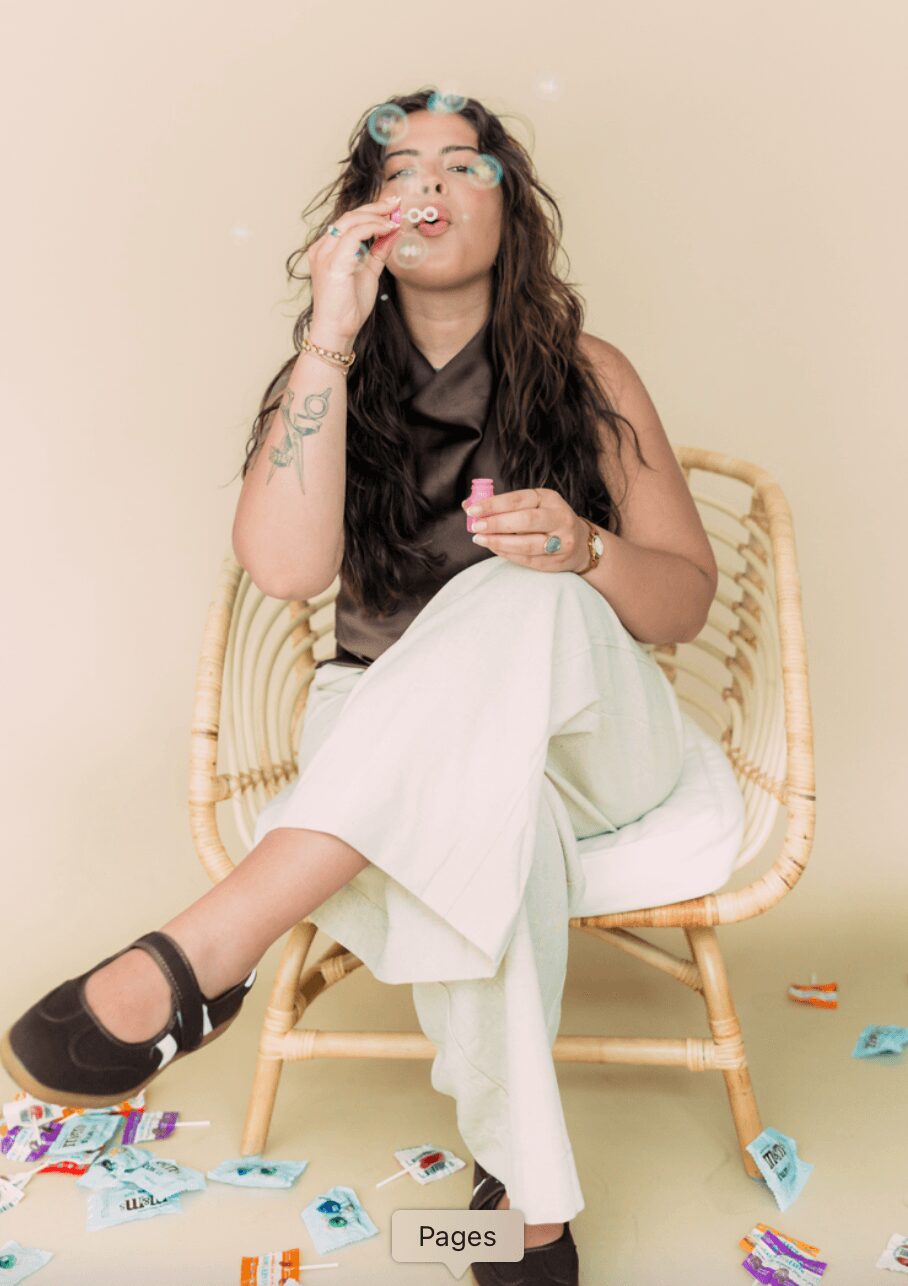 Woman with long dark hair sitting on a wicker chair, smoking, wearing a black top and white pants, with scattered objects on the floor.