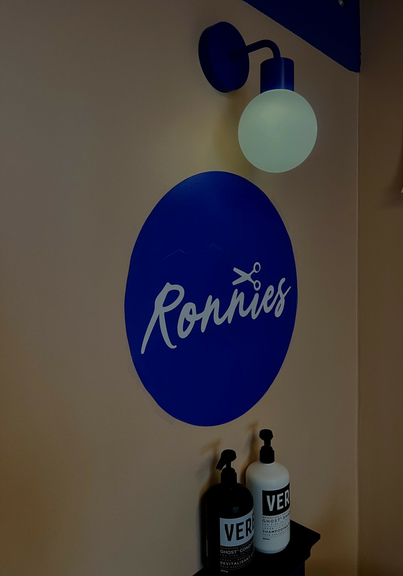 Wall sign with blue circle and white text 'Ronnies', two black and white bottles below, wall light above, beige wall background.