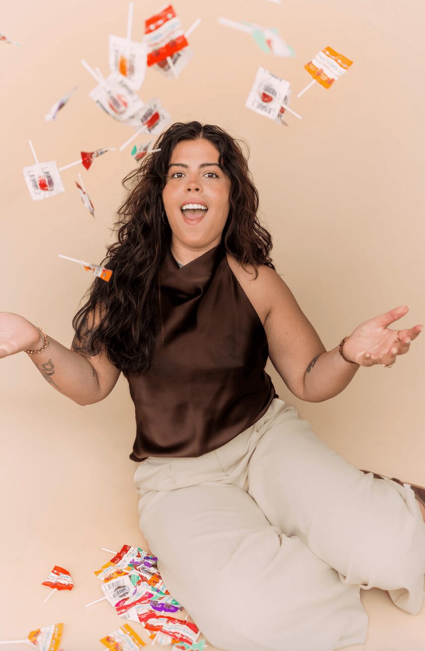 Woman with long curly hair sitting against a beige wall, smiling with arms outstretched, colorful confetti falling around her.