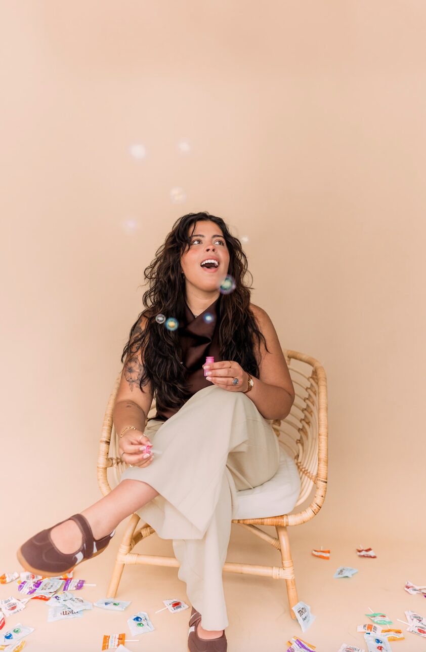 Woman with long dark hair sitting on a chair, surrounded by colorful confetti and bubbles, against a beige background.