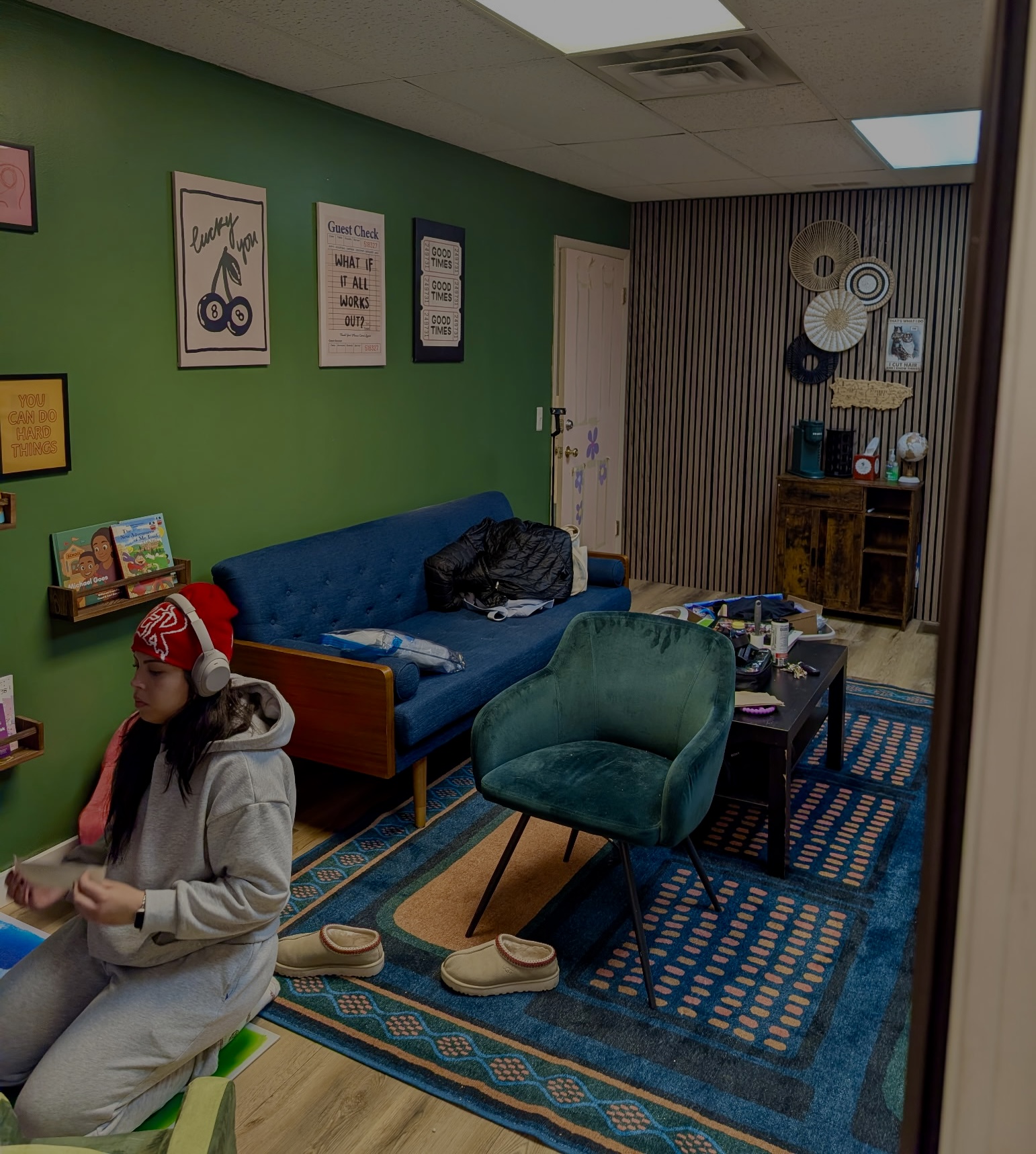 Person sitting on a green chair in a colorful room with a blue sofa, patterned rug, and wall decorations.