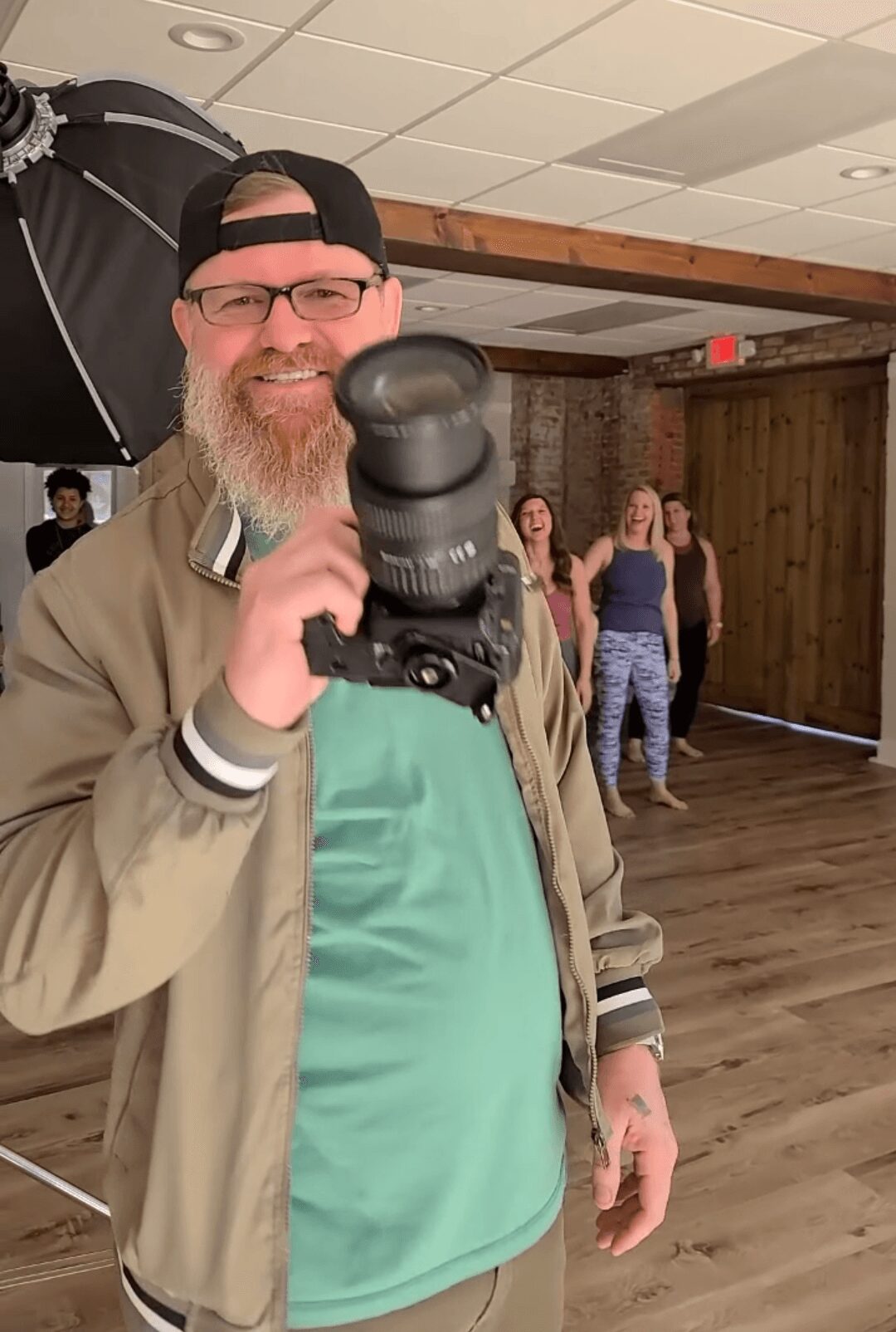 Man with glasses and a beard holding a camera, smiling indoors with people in the background.