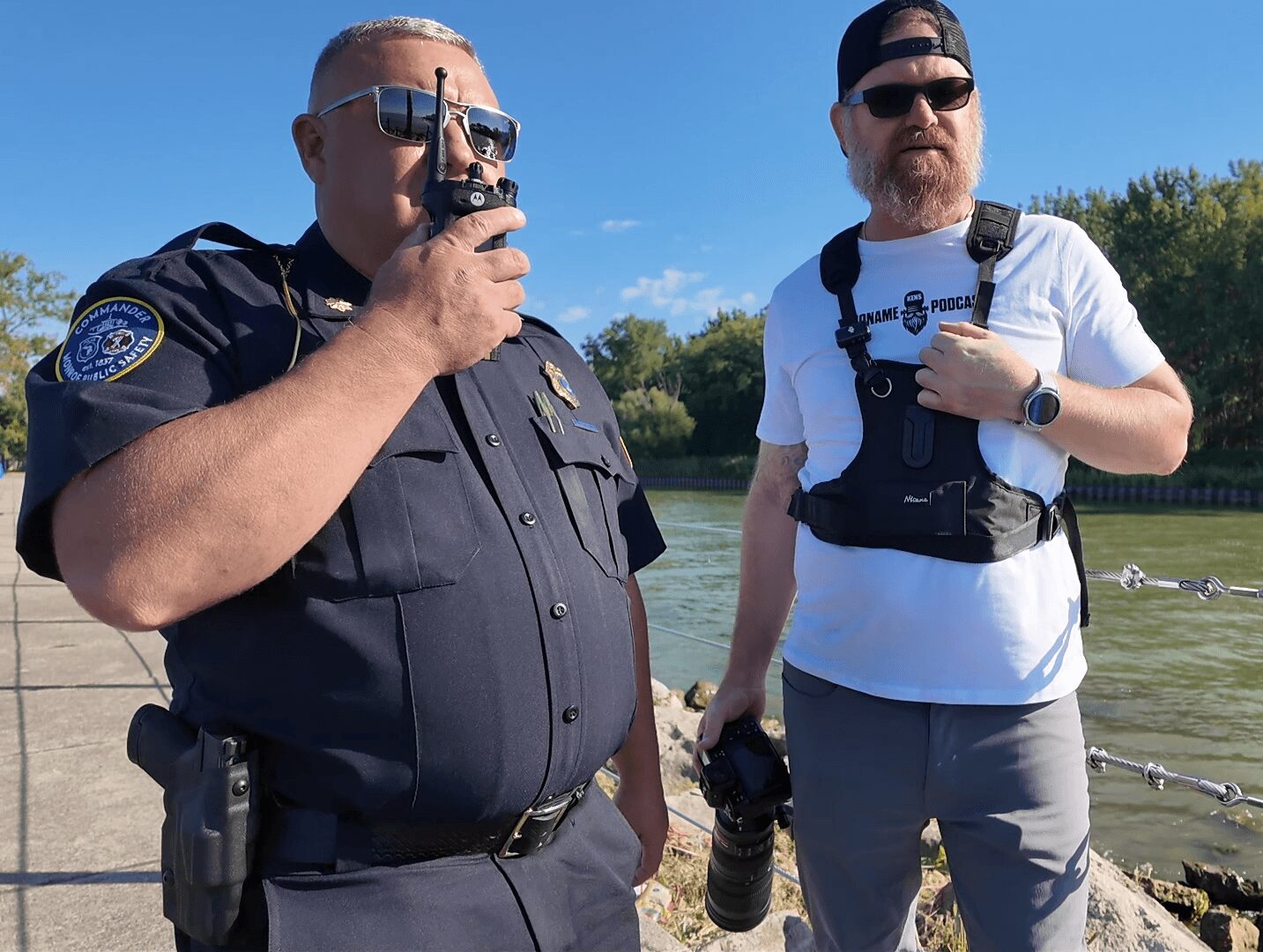 Two men outdoors, one in police uniform drinking from a bottle, the other in casual clothes holding a camera, with trees and water in background.