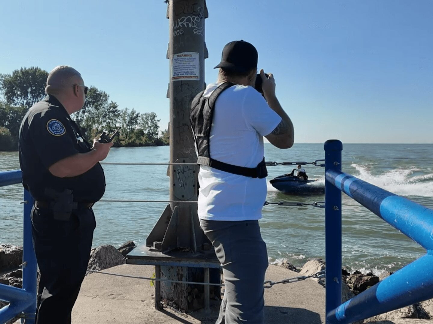 Two men near water, one in a police uniform and the other taking a photo, with a boat in the background.