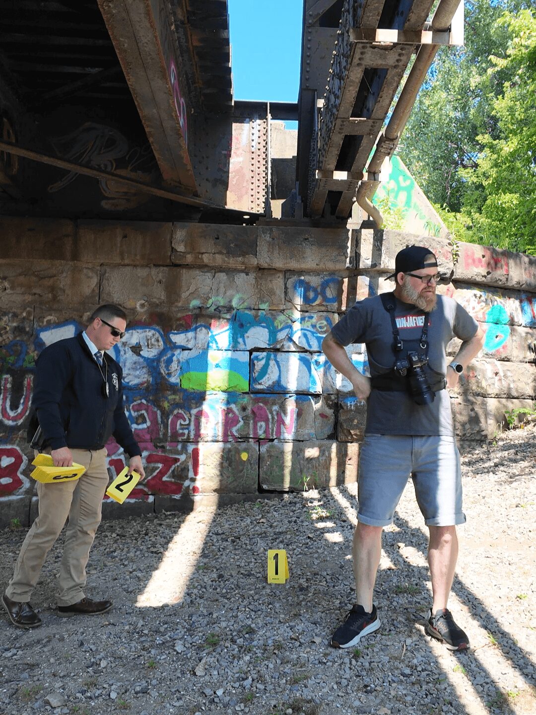 Two men standing outdoors near a graffiti-covered wall and a bridge, one with a camera, the other holding a yellow evidence marker.