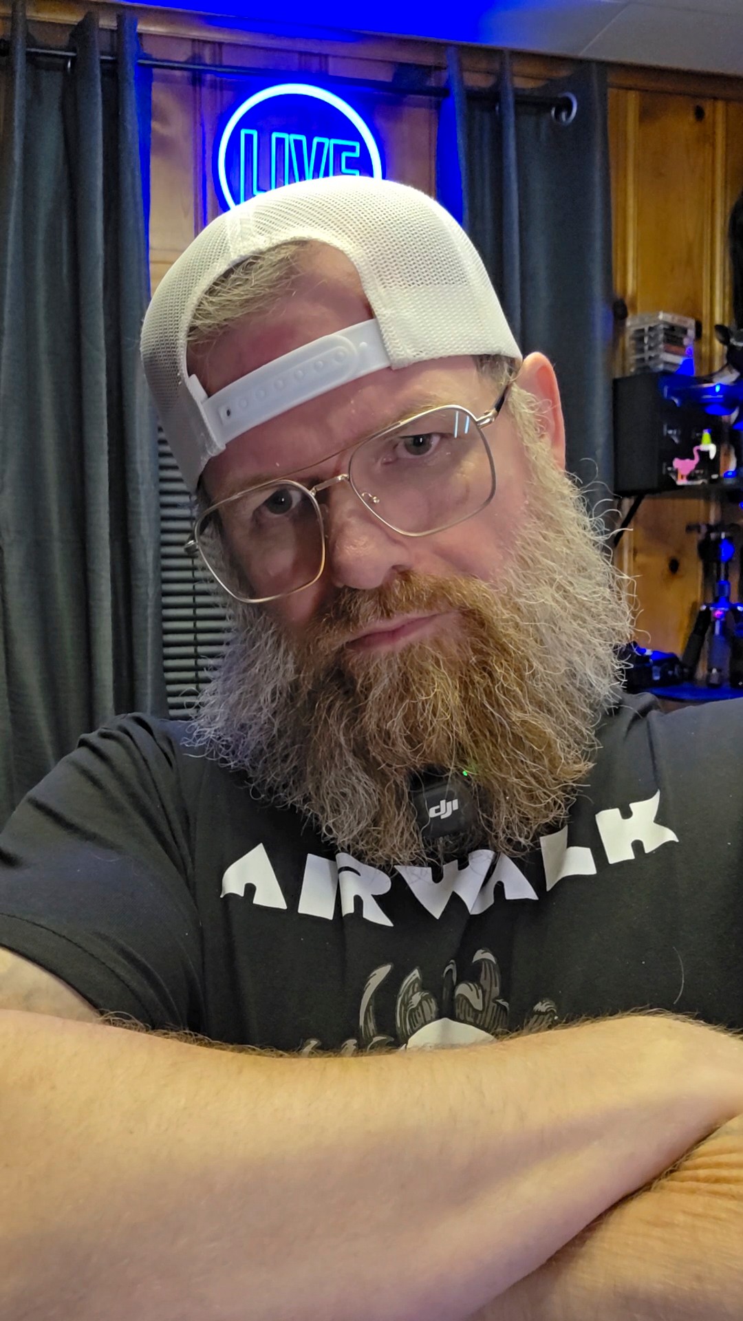 Man with glasses and a beard wearing a white cap, sitting indoors with a neon sign in the background.