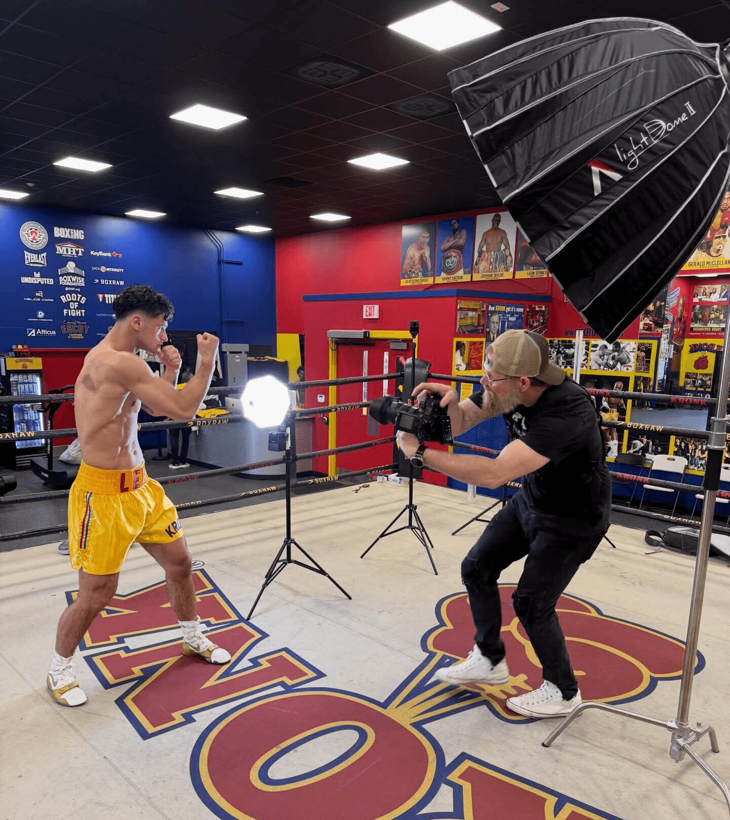 Boxer in yellow shorts and gloves training in a gym with a photographer filming him, professional lighting setup, boxing ring, and posters in background.