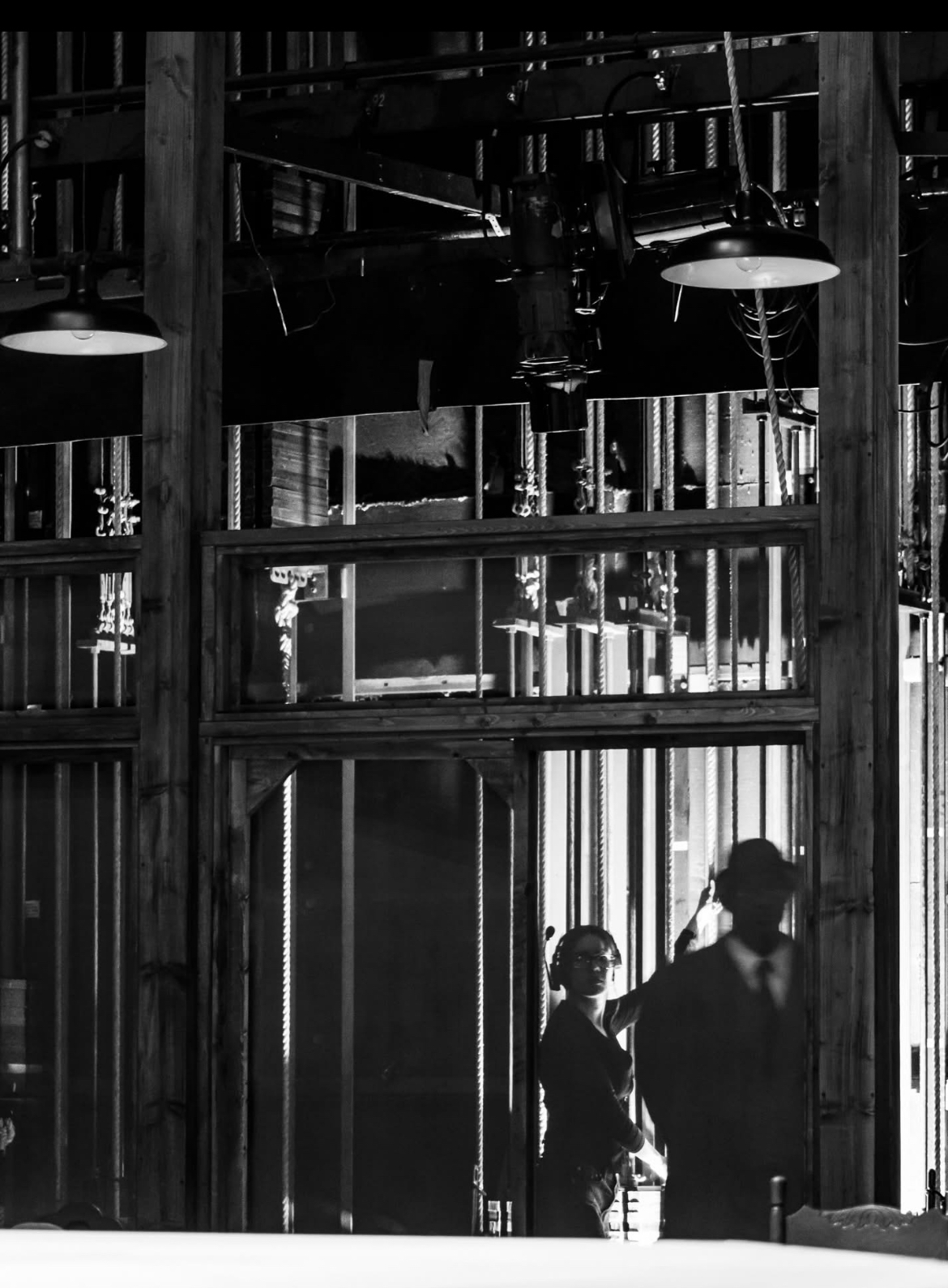 Two people standing inside a dark industrial space with metal beams and hanging lights, viewed through a window or glass door.