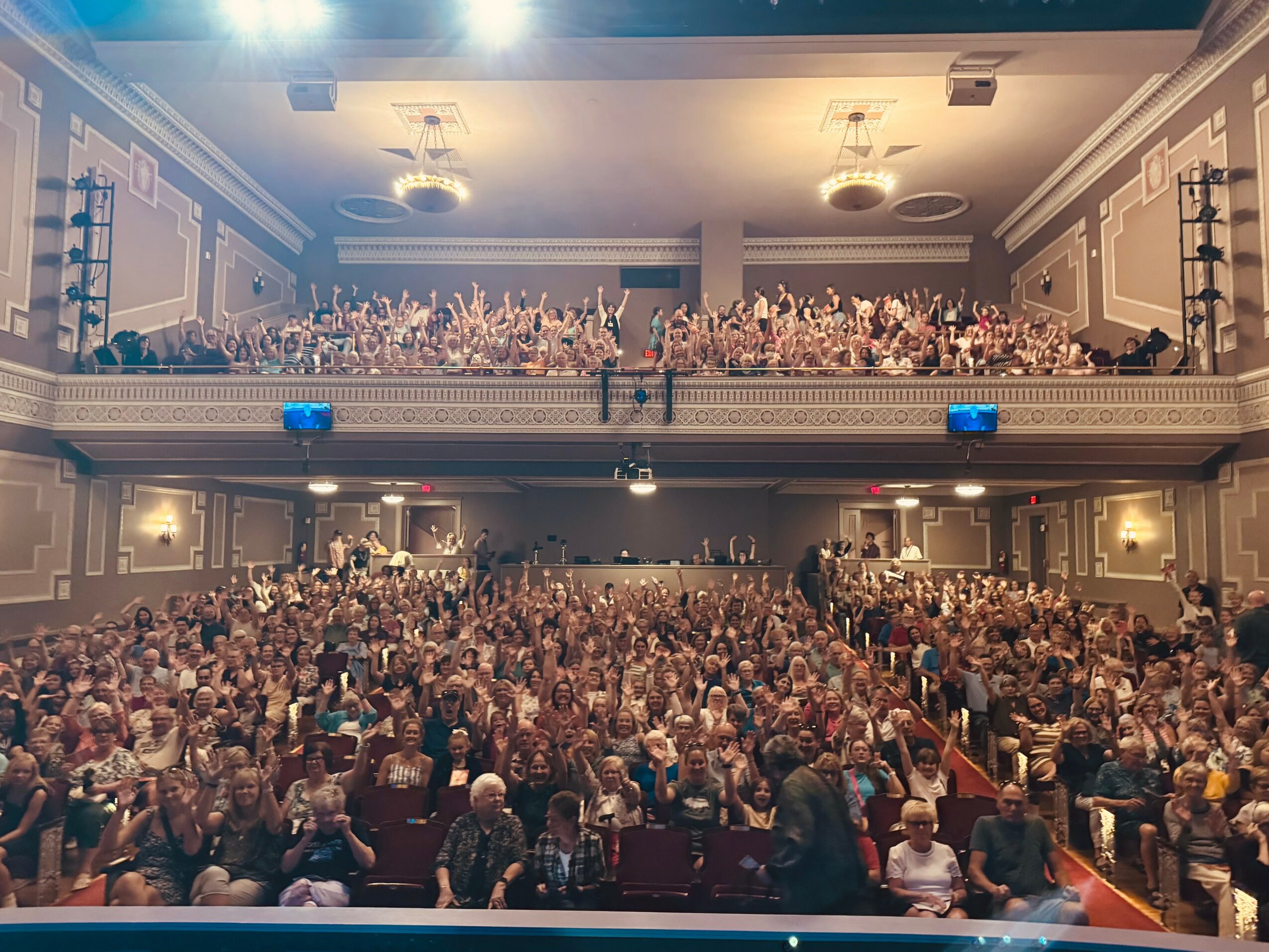 Large audience seated in a theater with ornate walls and balcony, facing stage, with some standing and talking.