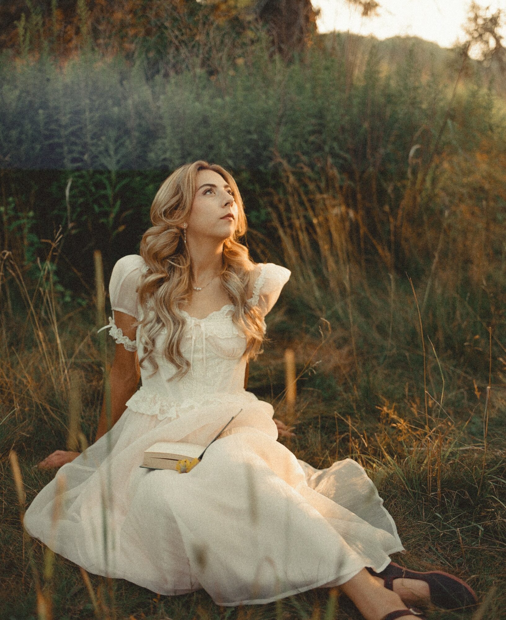 Young woman in a white dress sitting outdoors in a grassy field during sunset, looking upward with long wavy hair.